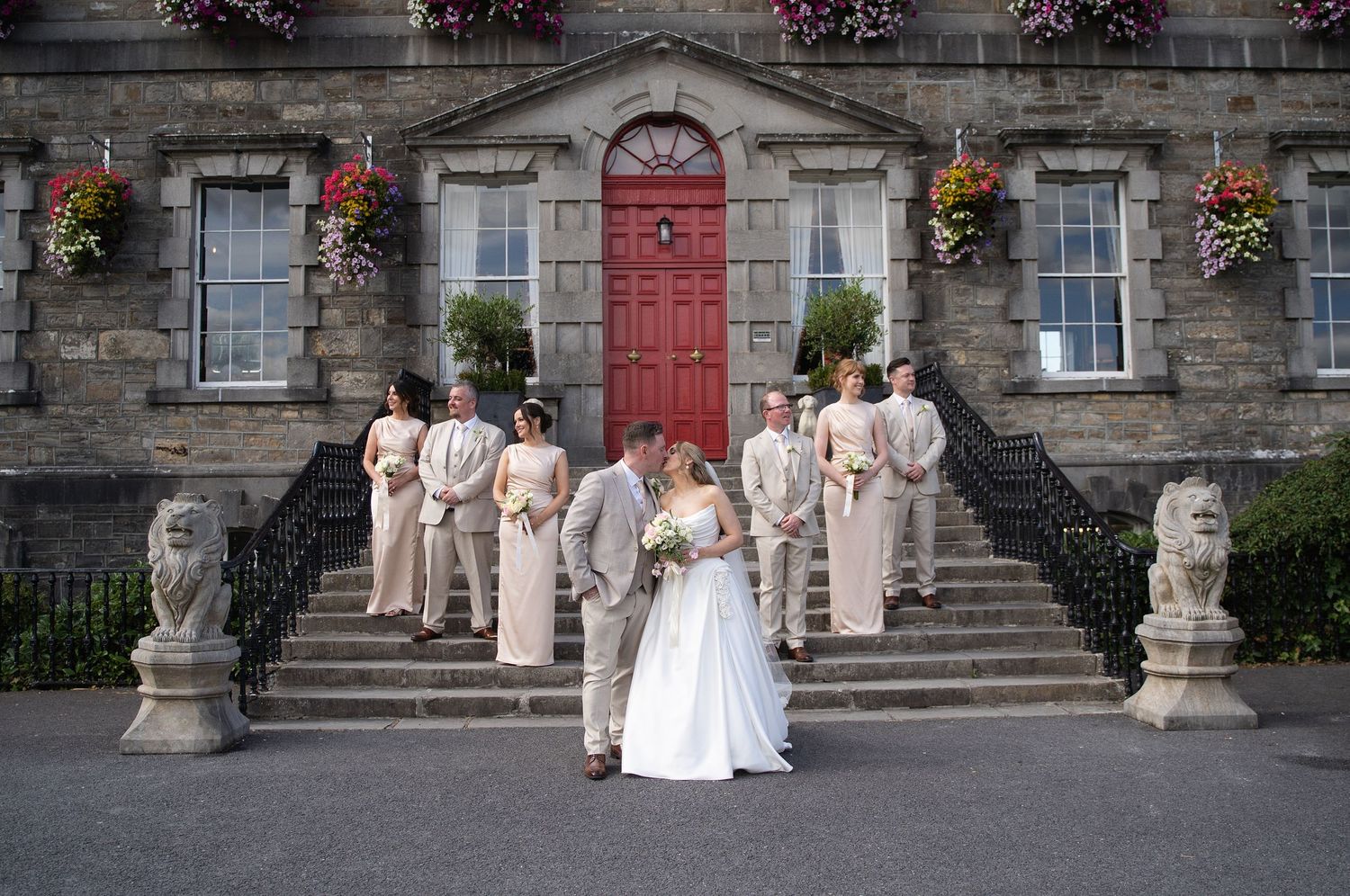 Wedding party poses on stone steps of elegant venue with red door, bride and groom kissing in foreground at Bellinter house.