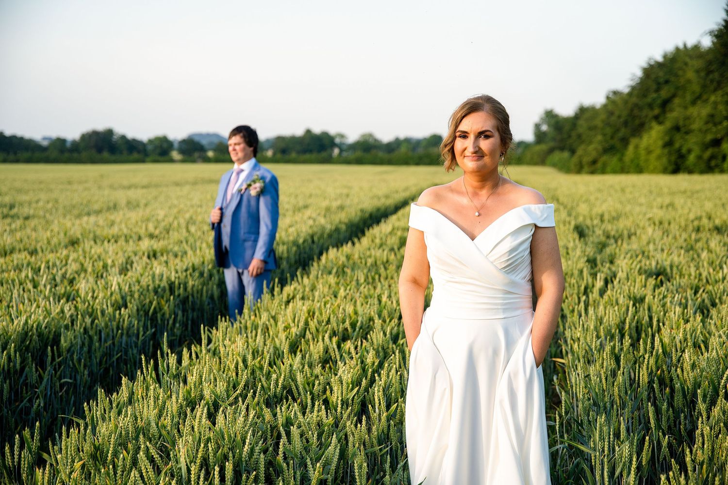 Bride in white off-shoulder gown stands in green field with groom in blue suit behind her on sunny wedding day.