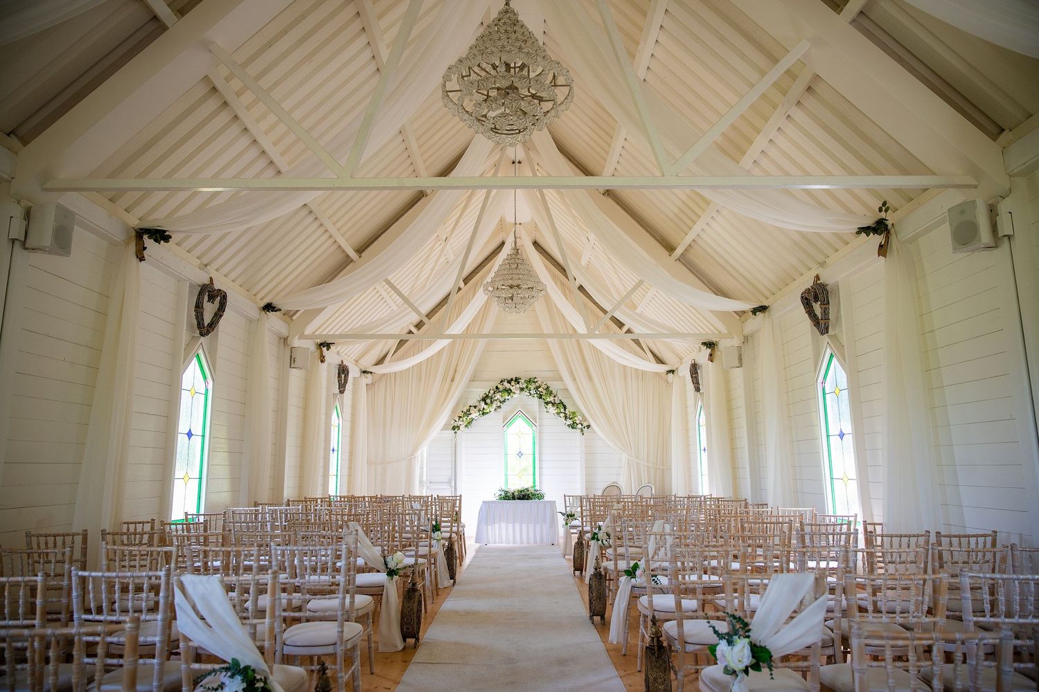 Elegant white chapel wedding venue with draped fabric ceiling, chandelier, wooden chairs, and floral arch altar.