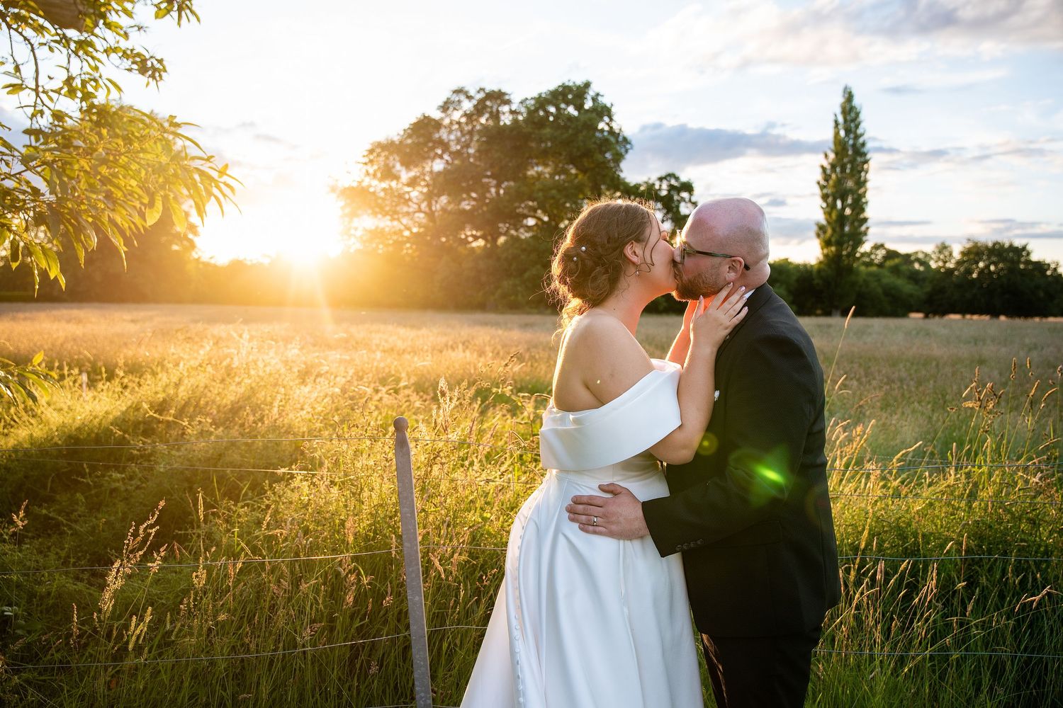 Bride and groom share a romantic sunset kiss in a golden field, with warm sunlight glowing behind lush green trees.