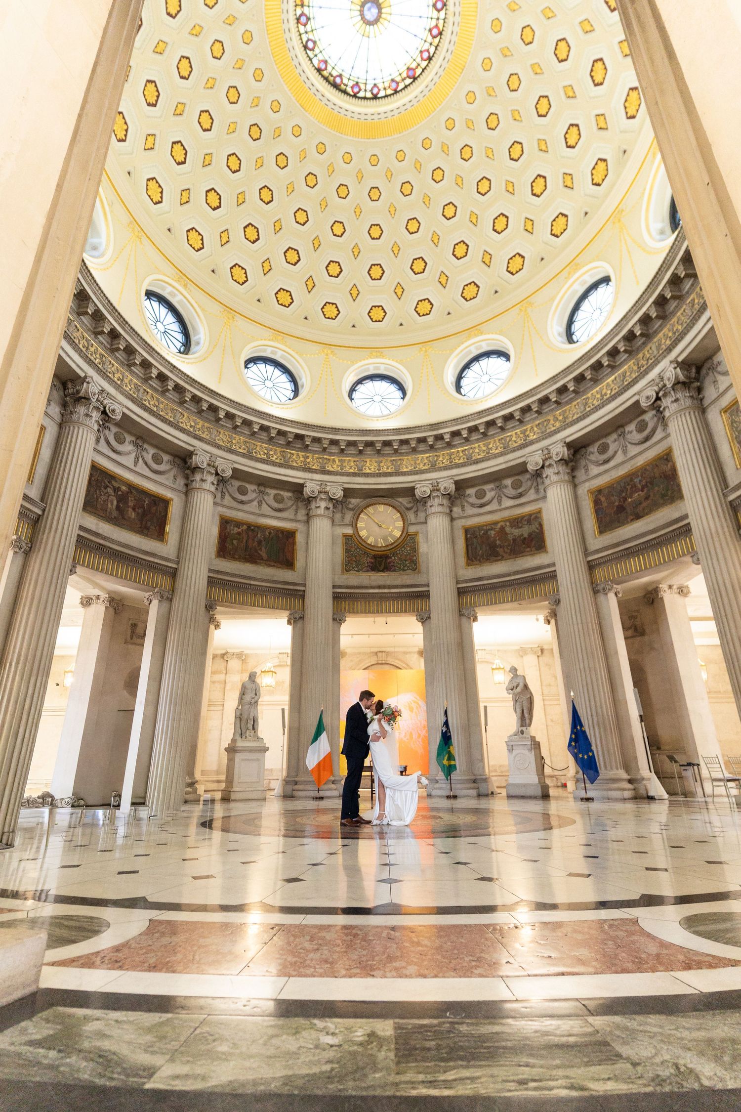 Couple embracing under ornate neoclassical dome with marble floors, columns, and Irish and EU flags in grand rotunda.