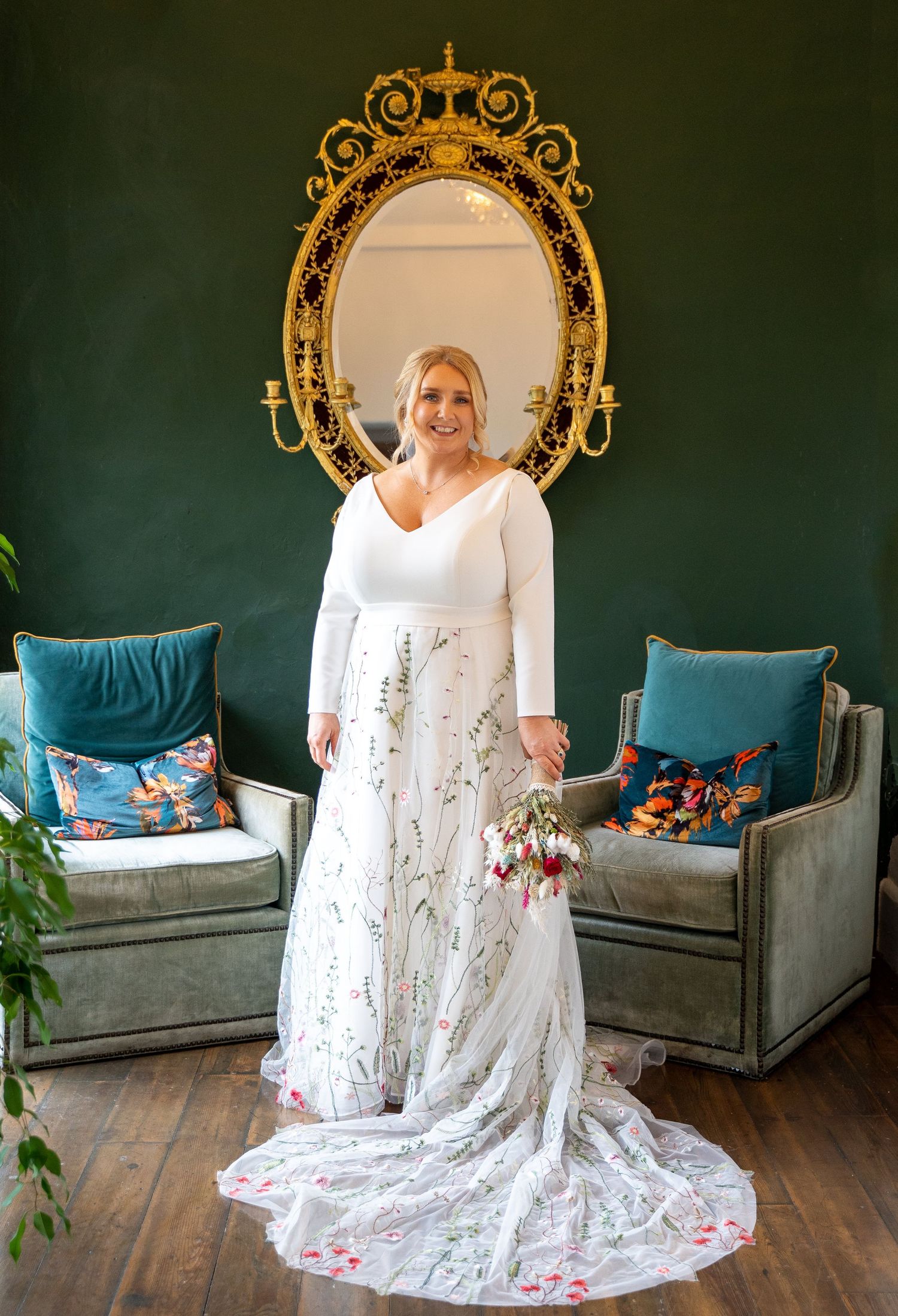 Bride in long-sleeve white floral wedding gown holding bouquet, standing before gold ornate mirror on dark green wall at Bellinter house.