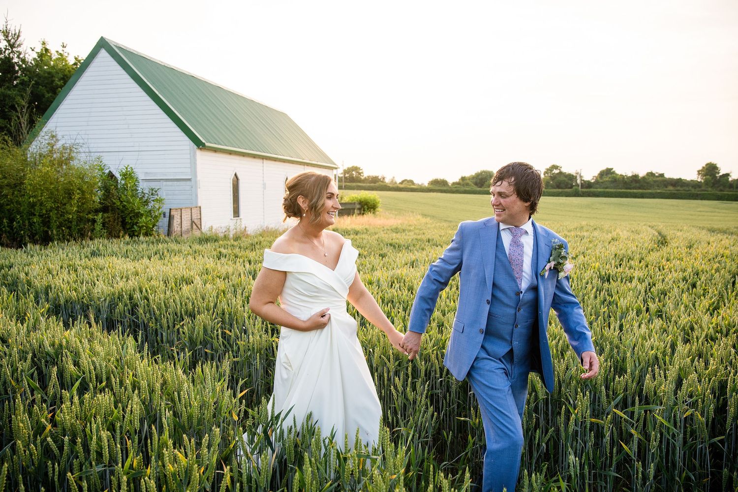 Bride and groom holding hands in lush green field at golden hour with white barn in background.