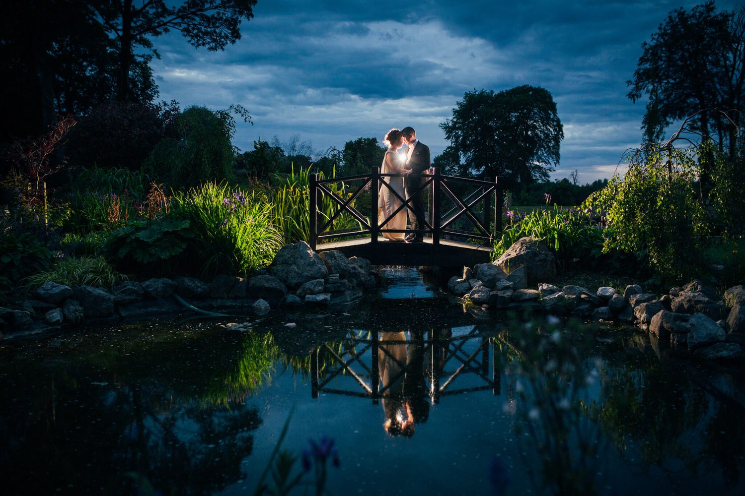 Bride and groom embrace on a lit wooden footbridge over a reflective pond at Darver Castle photo by AK wedding photography and videography.