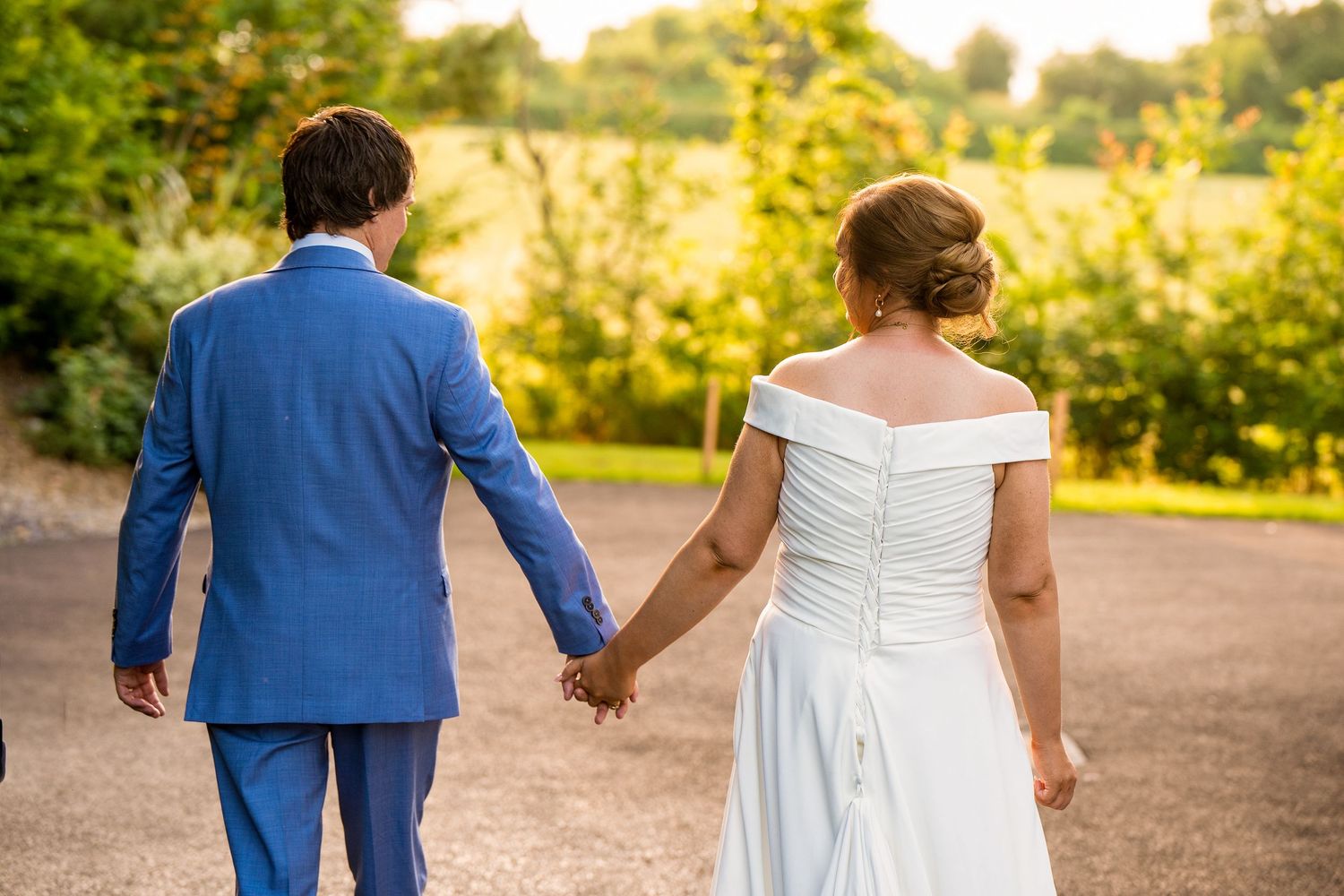 Bride and groom holding hands walking outdoors in blue suit and white off-shoulder wedding dress.