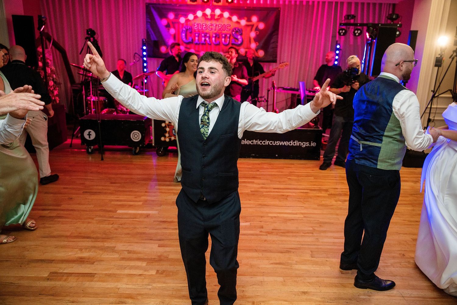 A man in a vest and tie dances with arms outstretched at a lively wedding reception with a band performing on stage.