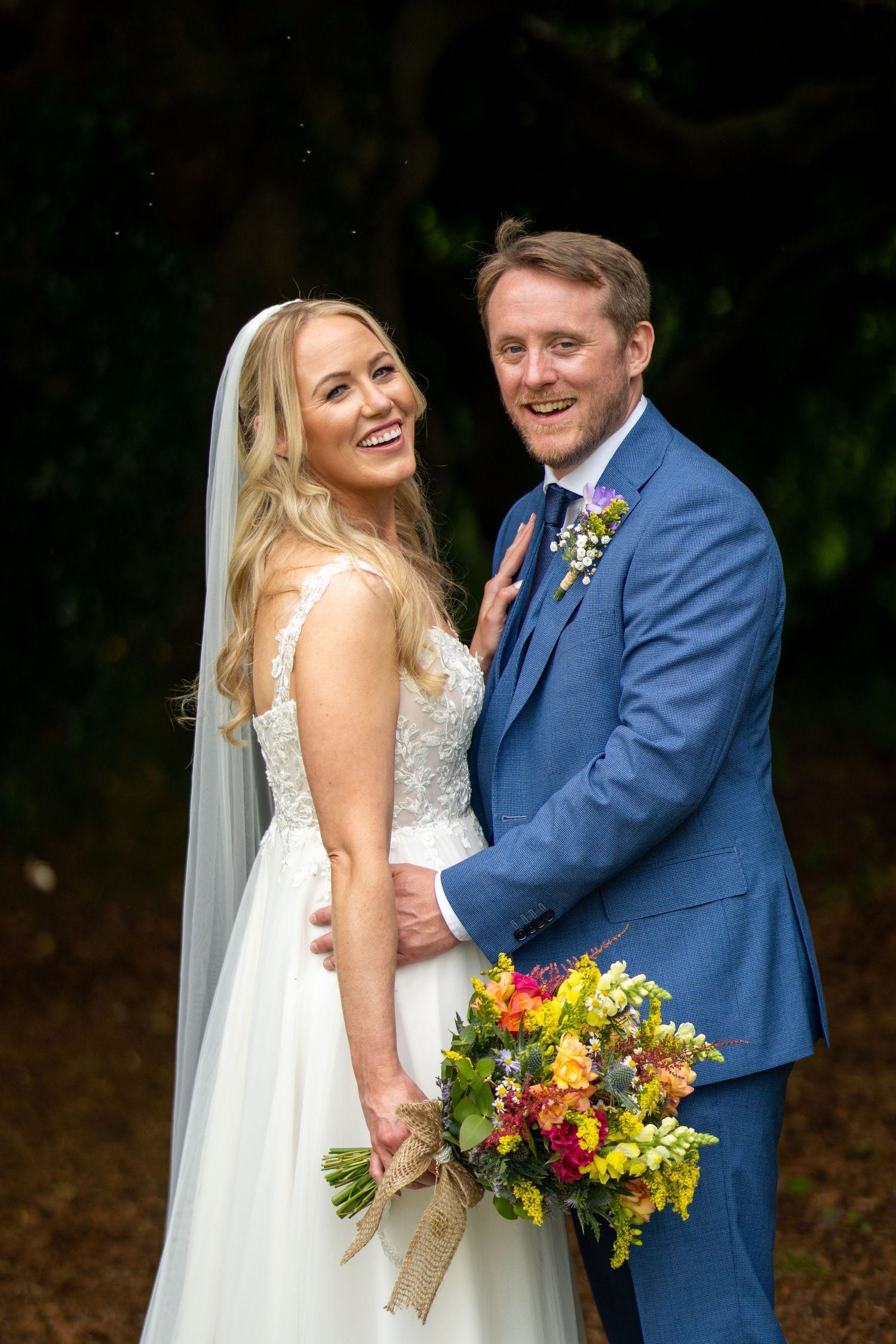 Smiling bride in white gown and veil holds colorful bouquet with groom in blue suit outdoors at Bellinter house.