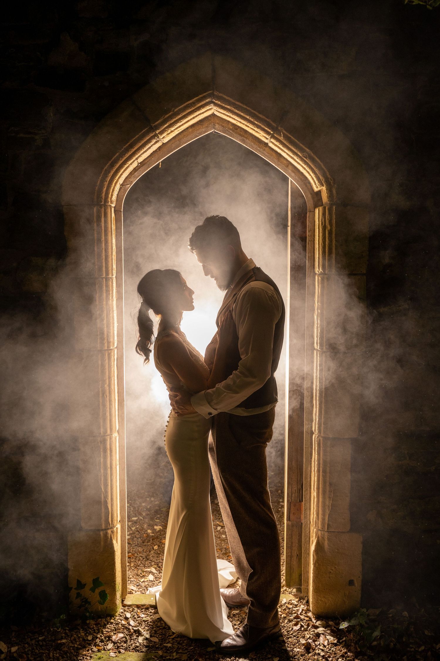 Silhouetted couple shares romantic moment in gothic archway with dramatic backlighting and atmospheric fog.