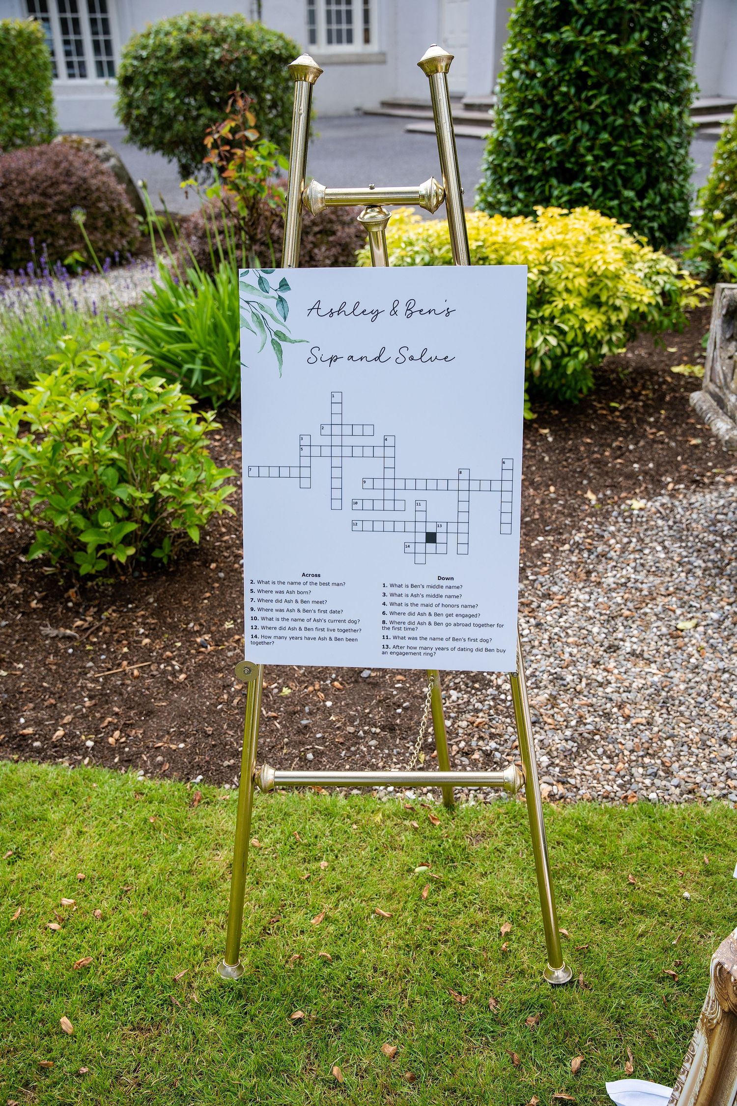 Gold easel displaying Ashley & Ben's Sip and Solve crossword puzzle wedding sign in a garden setting.
