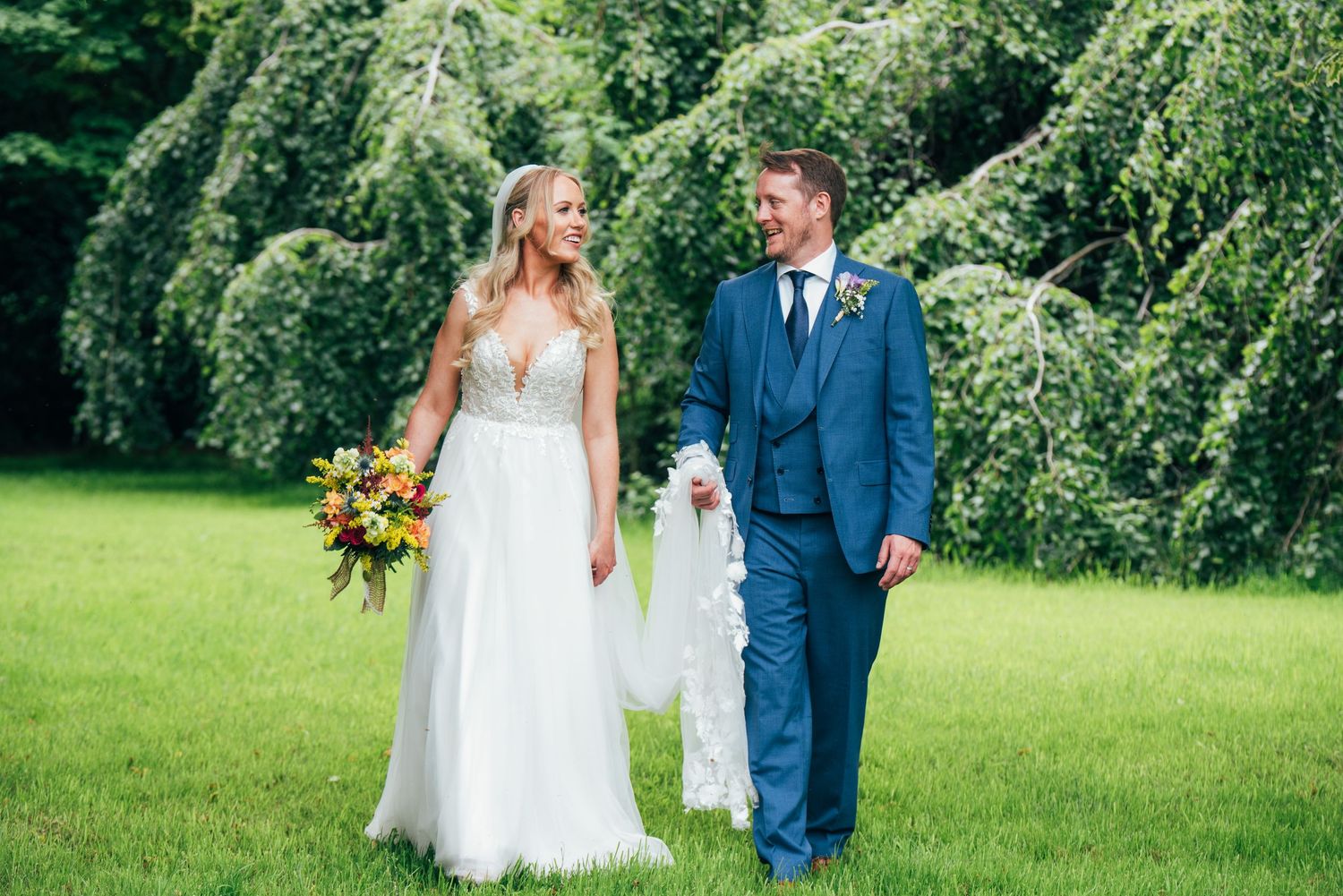 Bride in white gown and groom in blue suit walk hand in hand on green lawn with lush trees backdrop at Bellinter house.