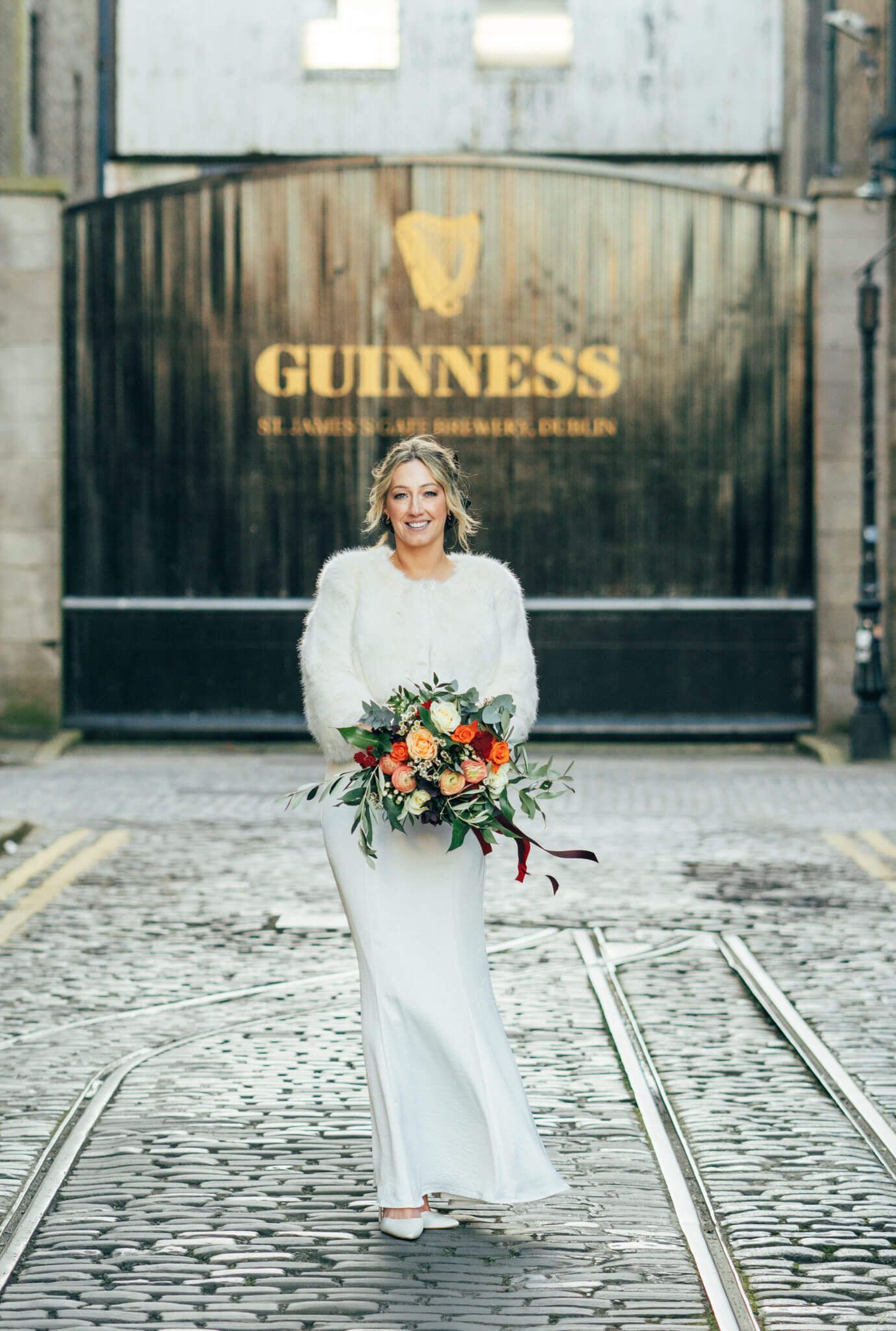 Bride in a white fur shrug  front of the Guinness Storehouse gates in Dublin photo by AK wedding photography and videography.