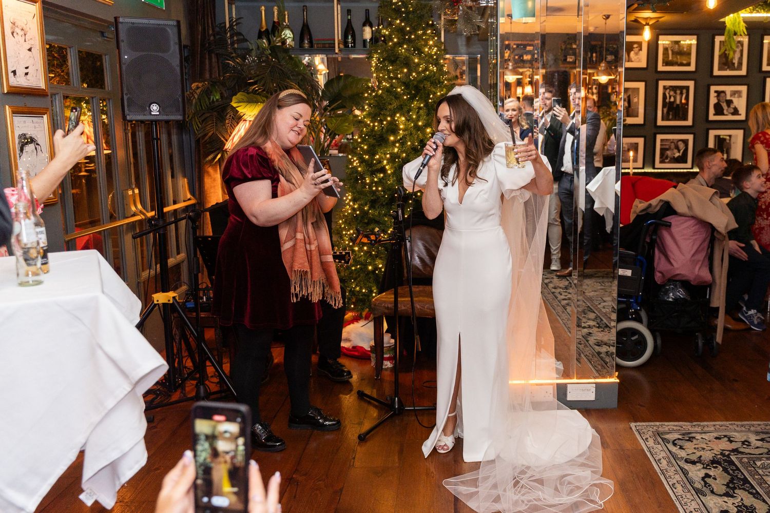 Bride in white gown and veil sings at wedding reception near Christmas tree while guest in red dress records on phone.