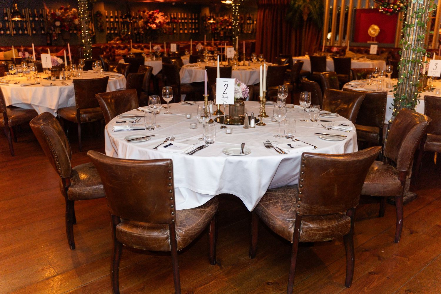 Elegant round dining tables set with white linens, candles, and glassware in a warmly lit restaurant.