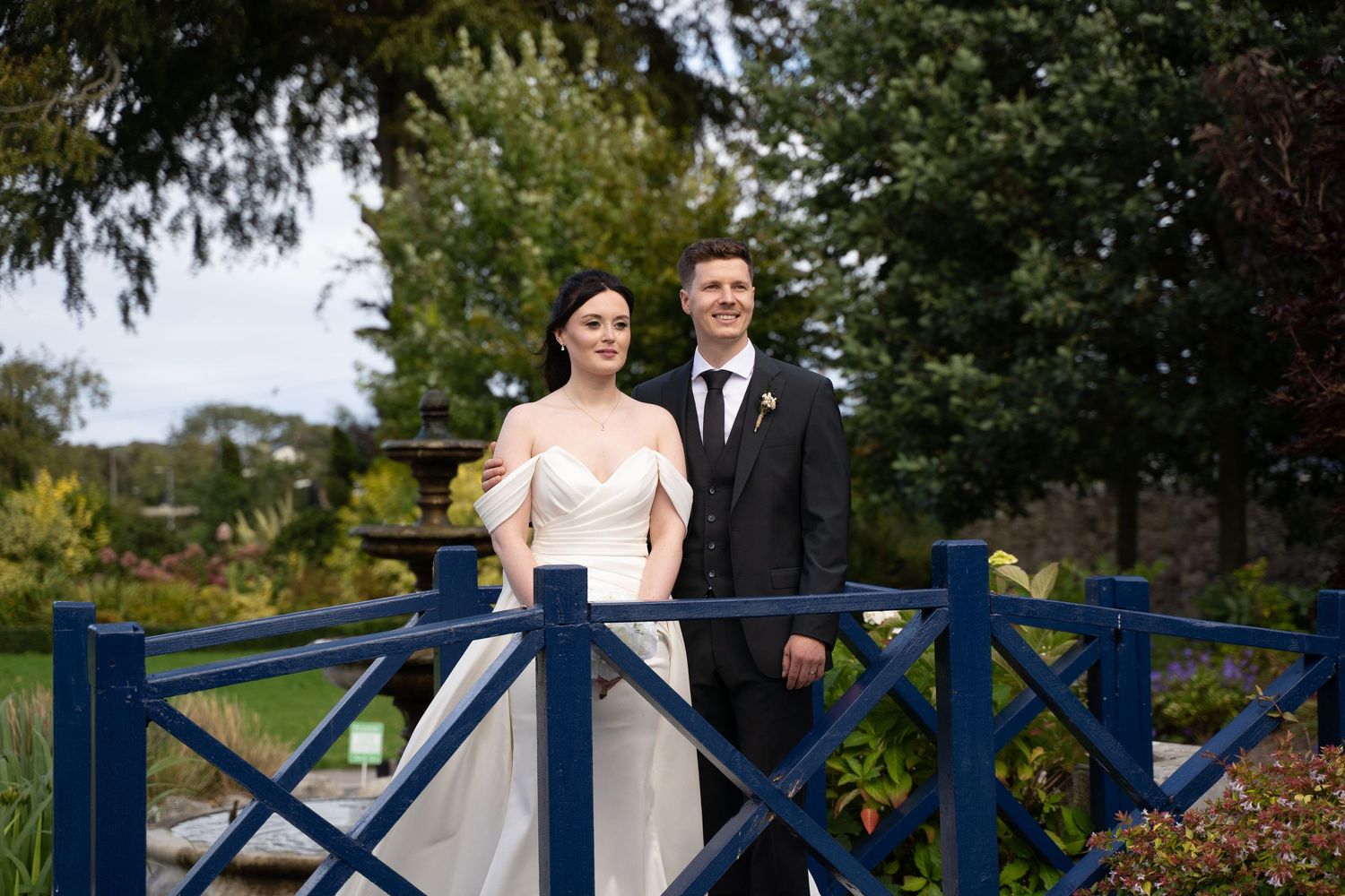 A couple in wedding attire stands together on a blue wooden bridge surrounded by lush greenery.