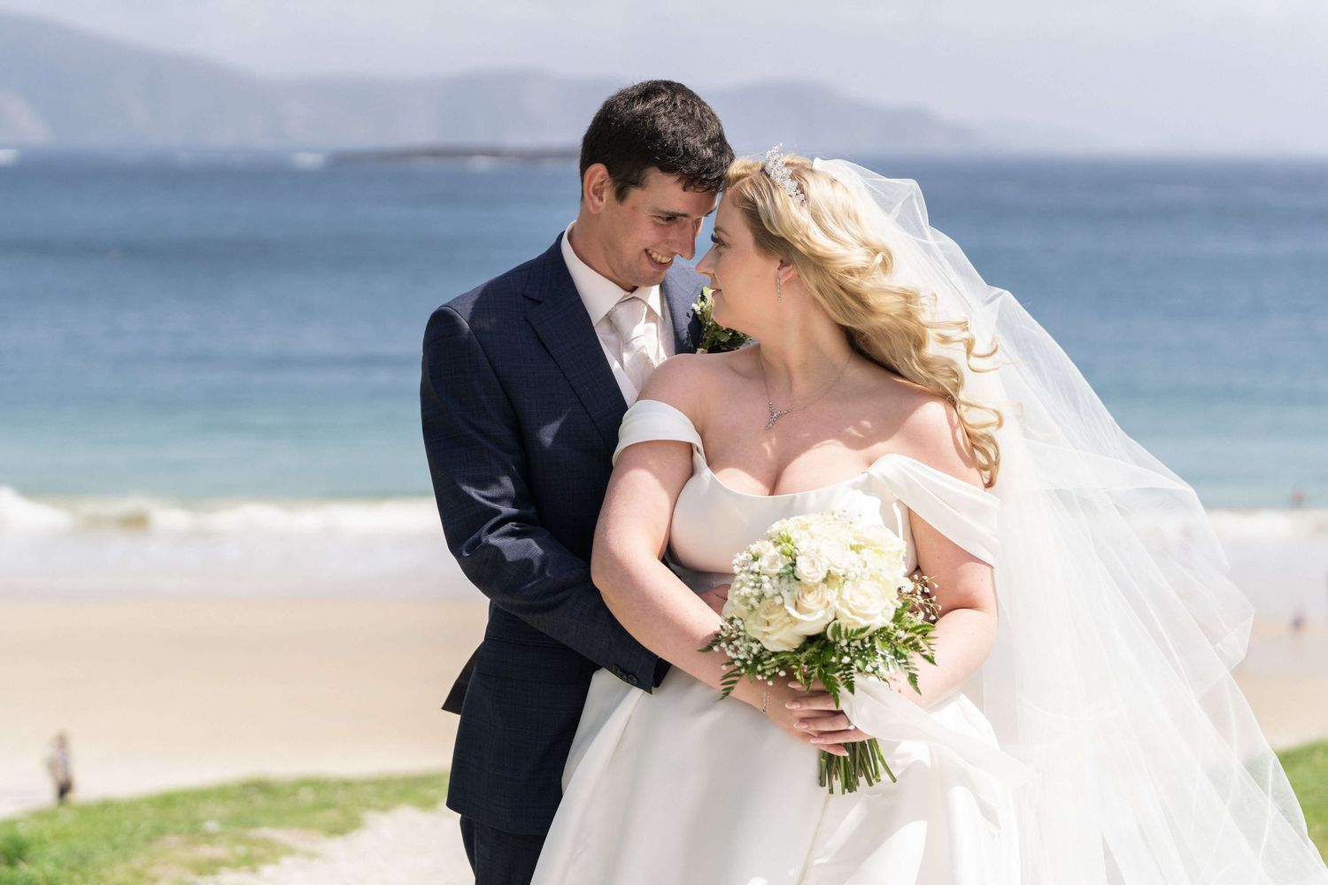 Bride in a white gown and veil holding a bouquet embraces her groom in a navy suit on a sandy beach with turquoise sea and mountains behind.