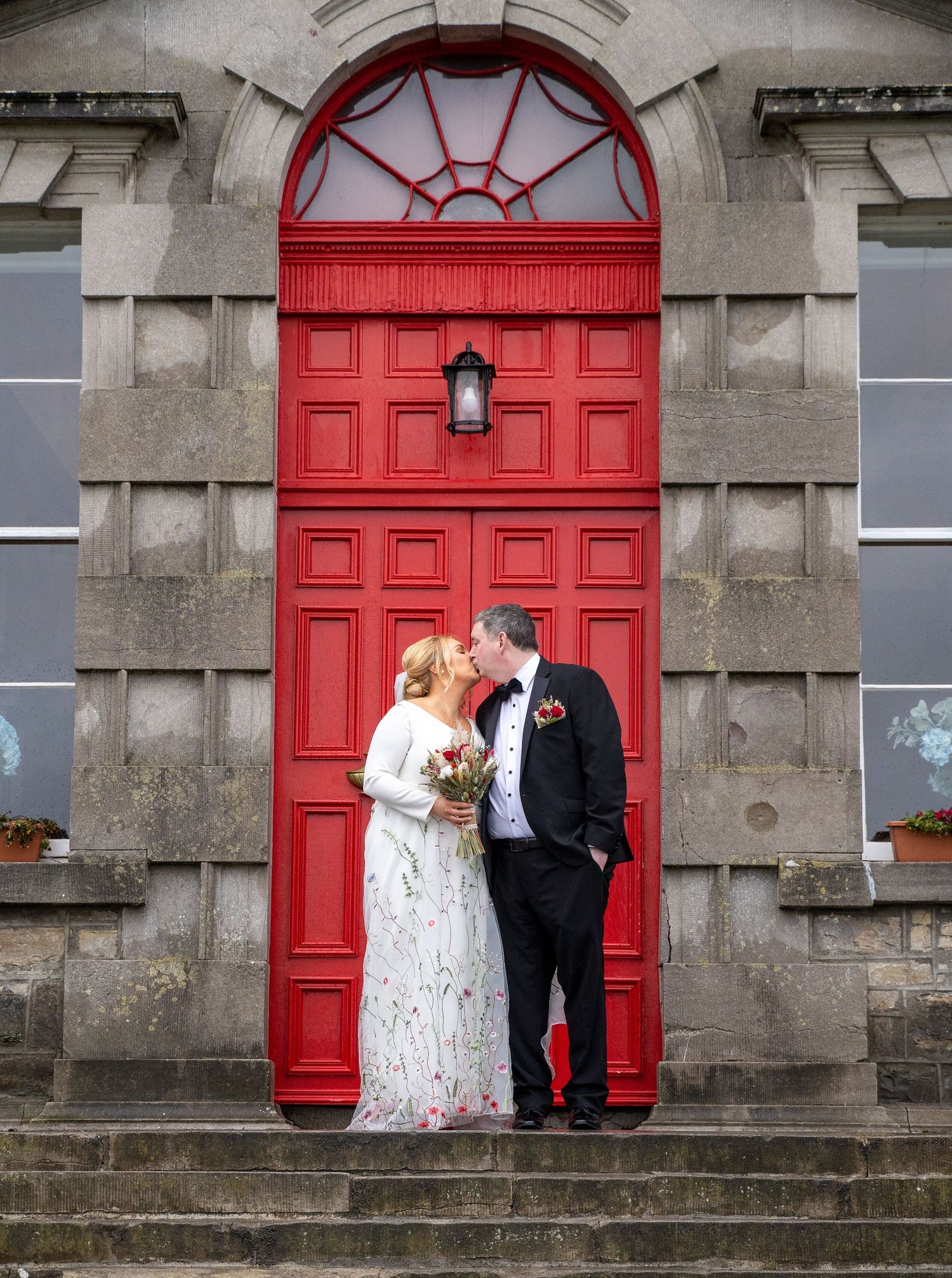 Couple sharing a kiss in wedding attire on steps before a grand red door of a historic stone building at Bellinter house.