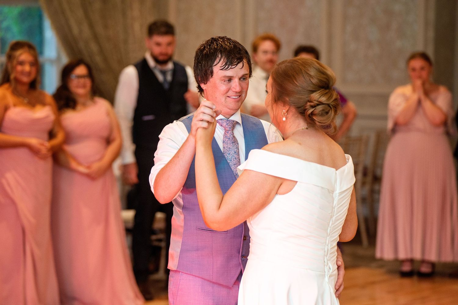 Bride and groom share their first dance at a wedding, with bridesmaids in pink dresses watching in the background.
