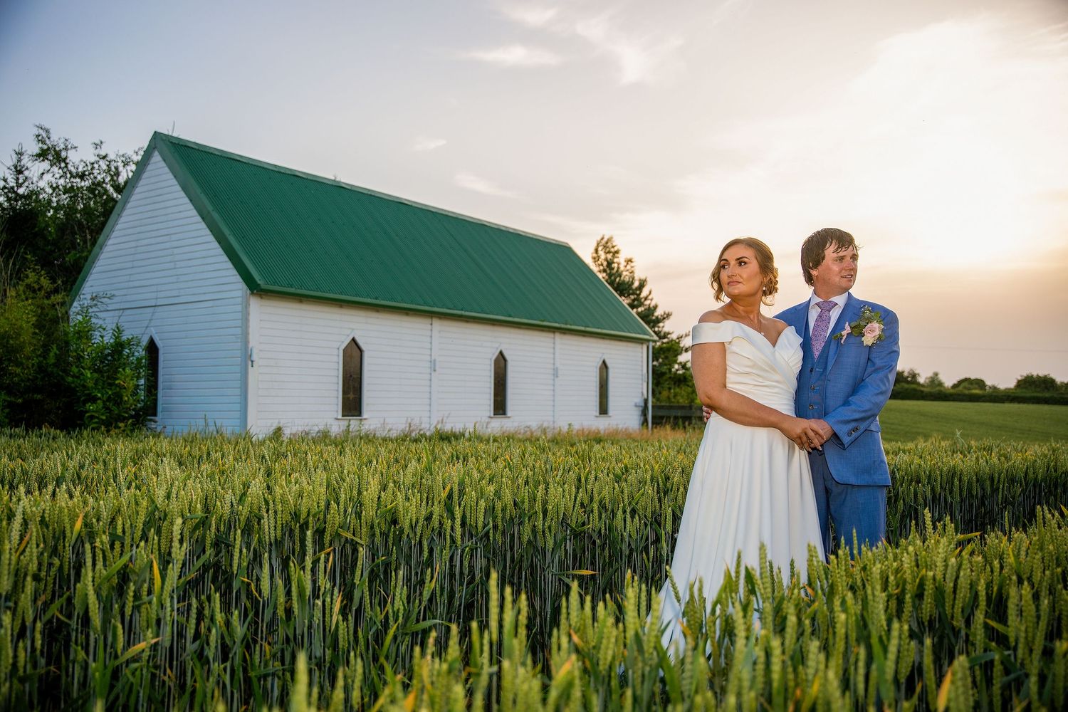 Bride and groom stand in a green wheat field before a white chapel with green roof at golden sunset.