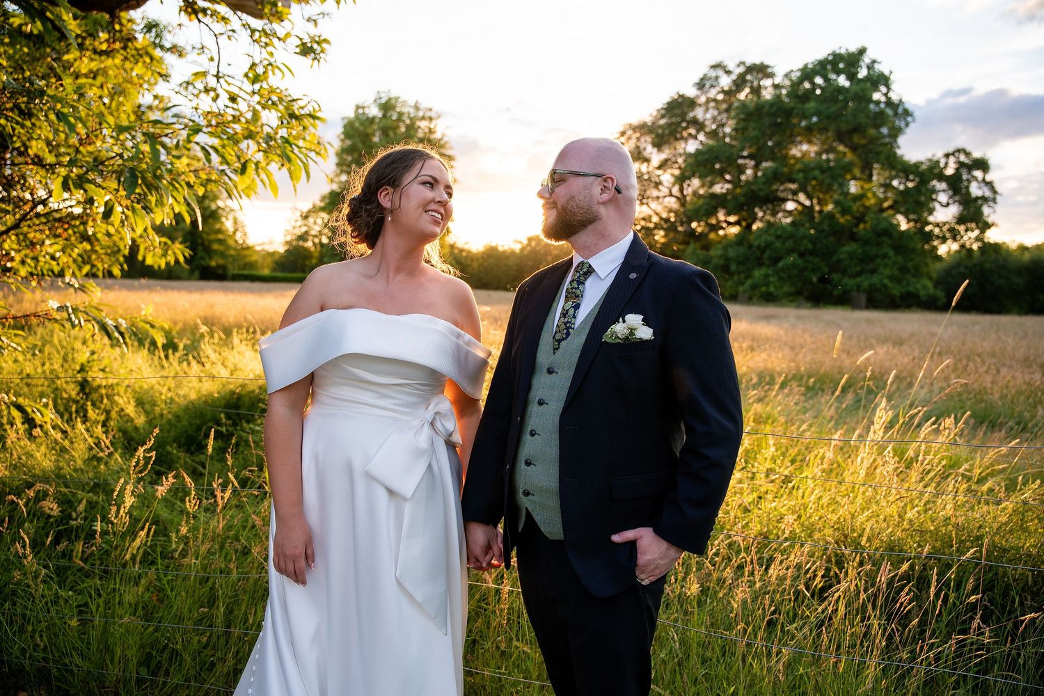Bride and groom holding hands at golden hour in a field, smiling at each other in wedding attire.
