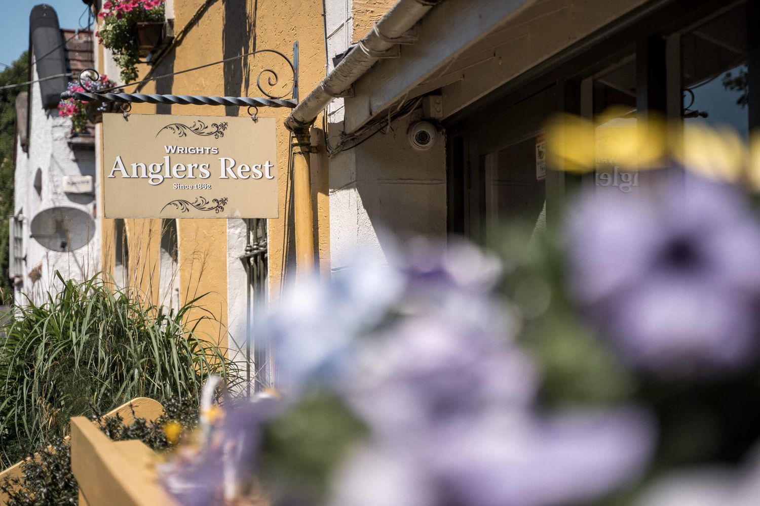 Anglers Rest pub sign on a sunlit stone building with hanging flower baskets and blurred purple flowers in the foreground.