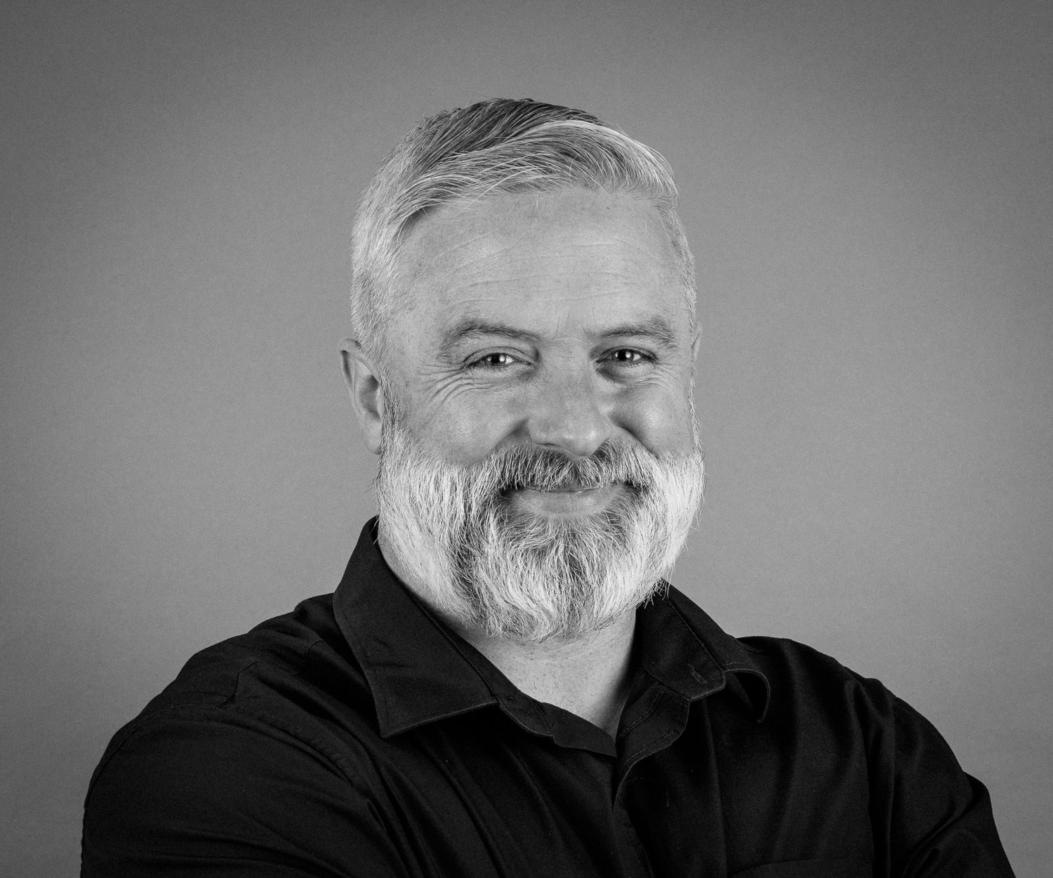 Black and white professional headshot of a smiling middle-aged man with gray hair and beard wearing a black shirt.