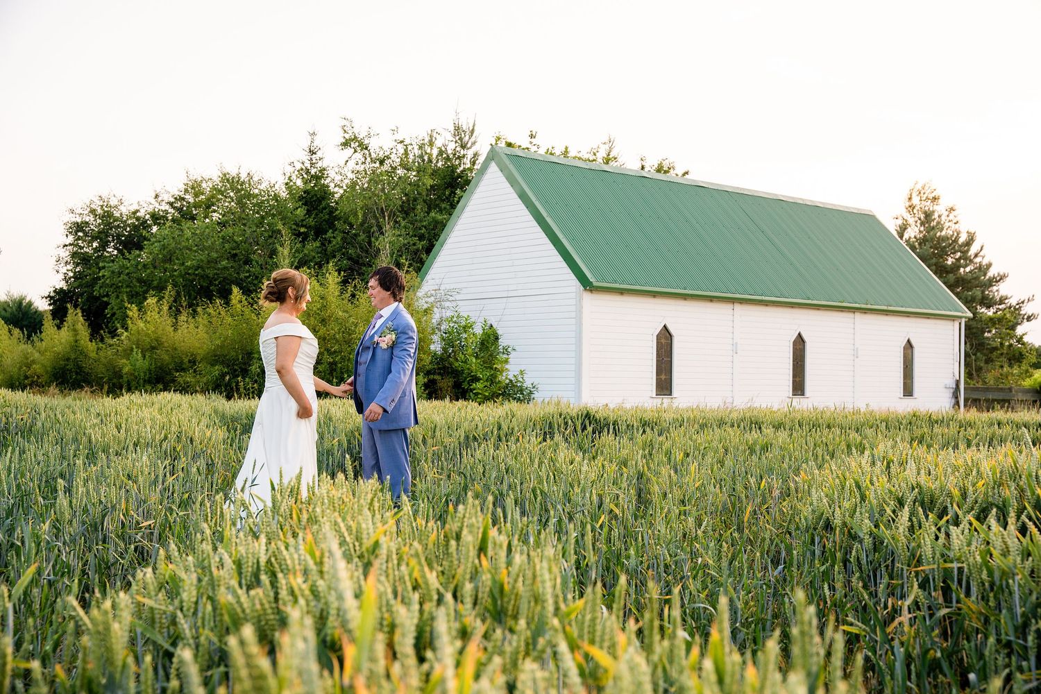 Bride and groom holding hands in a green wheat field with a white chapel and green roof in the background.
