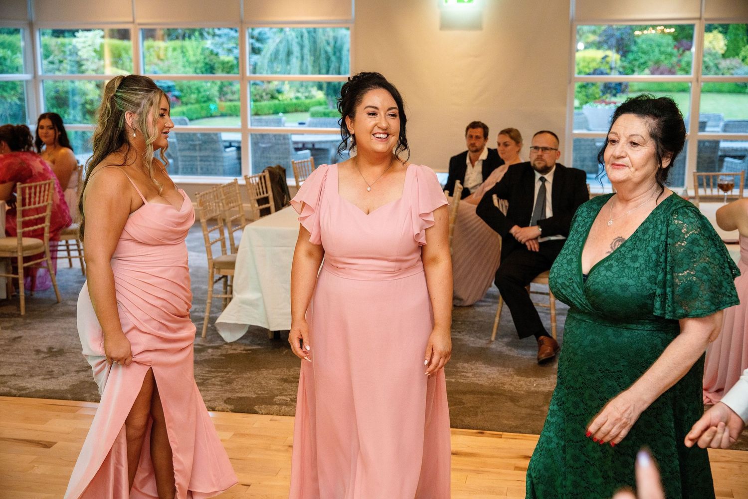 Three women in pink and green dresses laughing at a wedding reception with seated guests in the background.