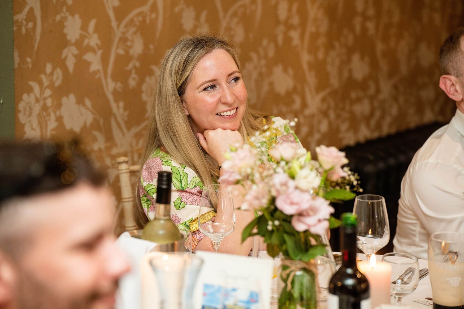 Smiling woman with long blonde hair sits at a dinner table beside a bouquet of pink flowers, with wine bottles and glasses nearby.