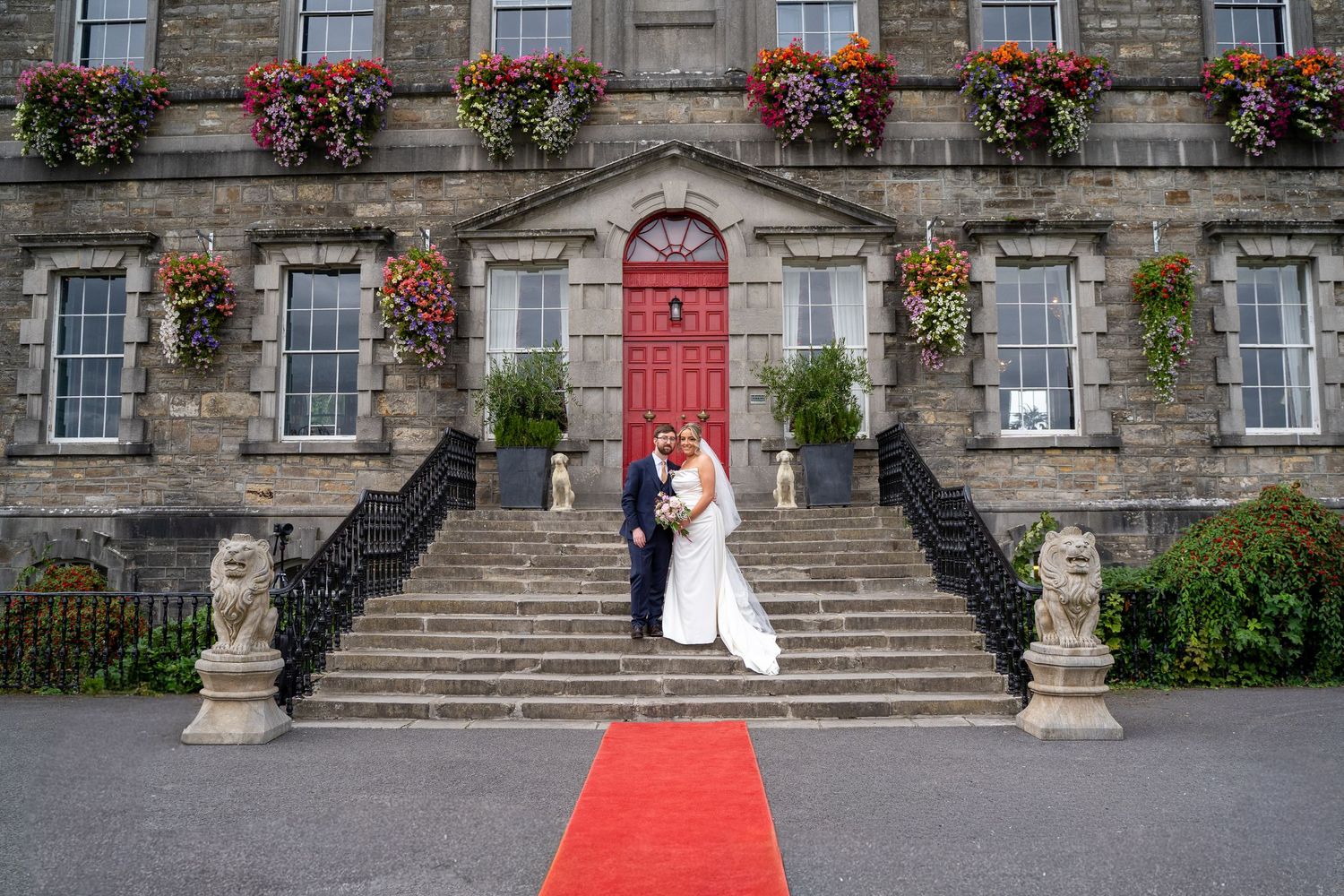 Bride and groom embrace on the stone steps of a historic manor with a red door, flower-filled window boxes, and a red carpet below.