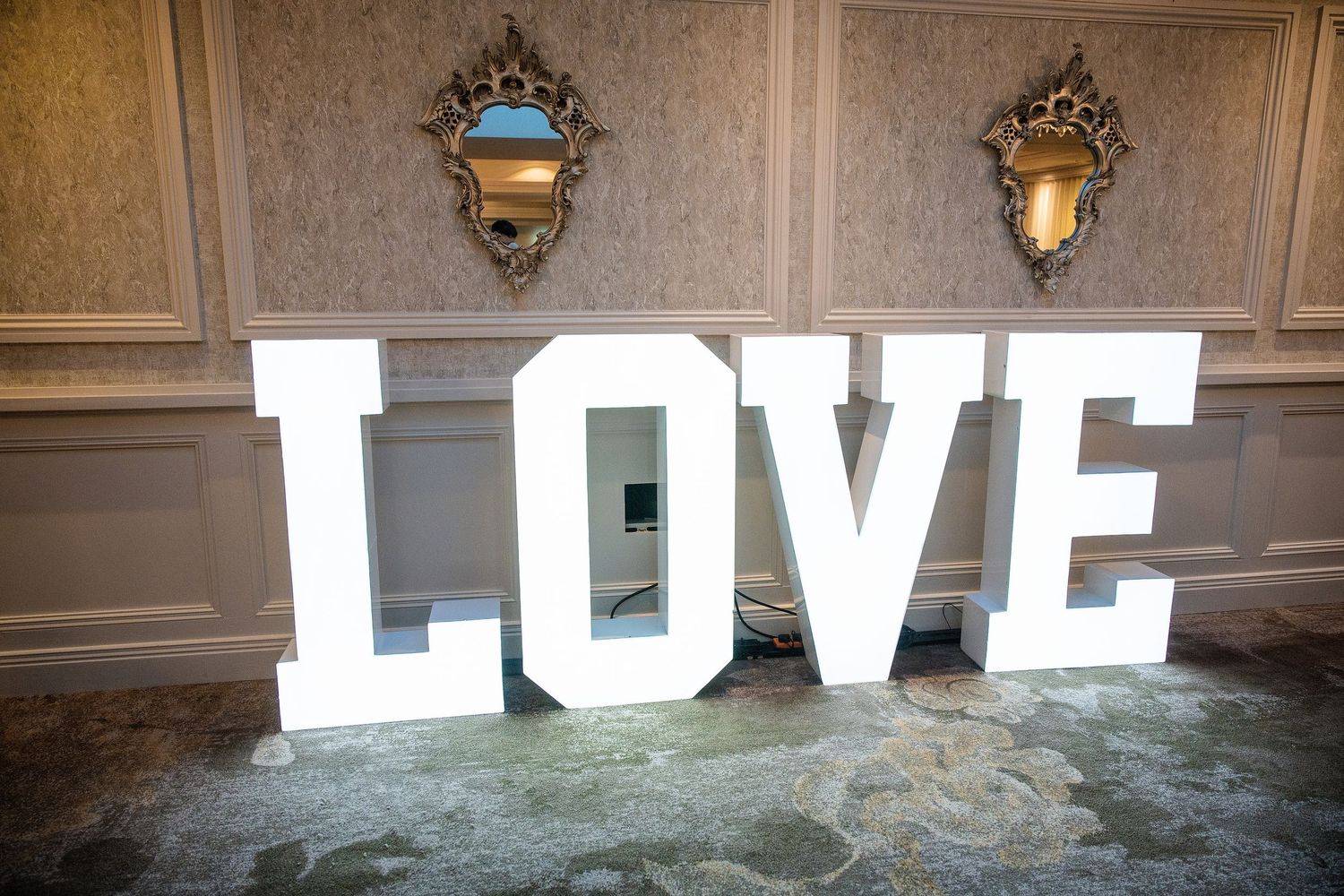 Large illuminated white LOVE letters display in an elegant wedding venue with ornate gold mirrors on wallpapered walls.
