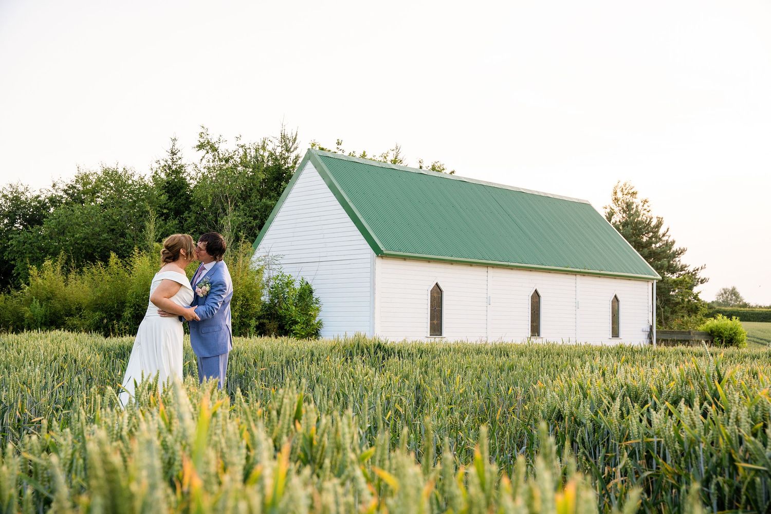 Couple embracing in a green wheat field in front of a white chapel with a green roof on their wedding day.