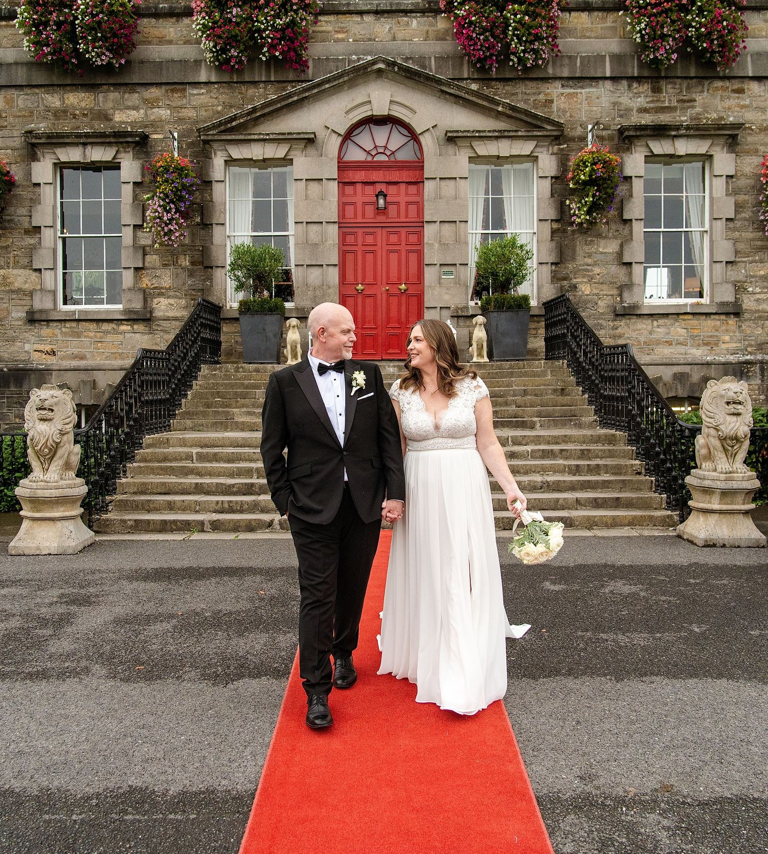 Bride and groom walking on red carpet outside elegant stone venue with red door and floral decorations at Bellinter house.