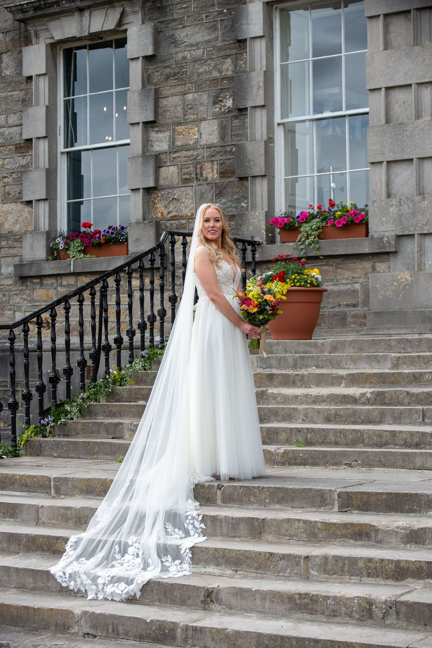 Bride in white lace gown with long veil holds colorful bouquet on stone steps Bellinter house.