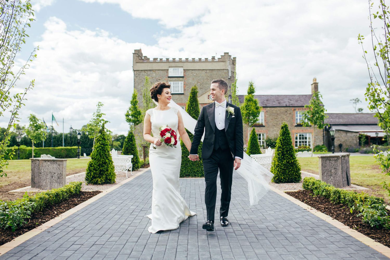 Bride holding a red bouquet and groom in a dark suit hold hands in front of Darver castle photo by AK wedding photography and videography.