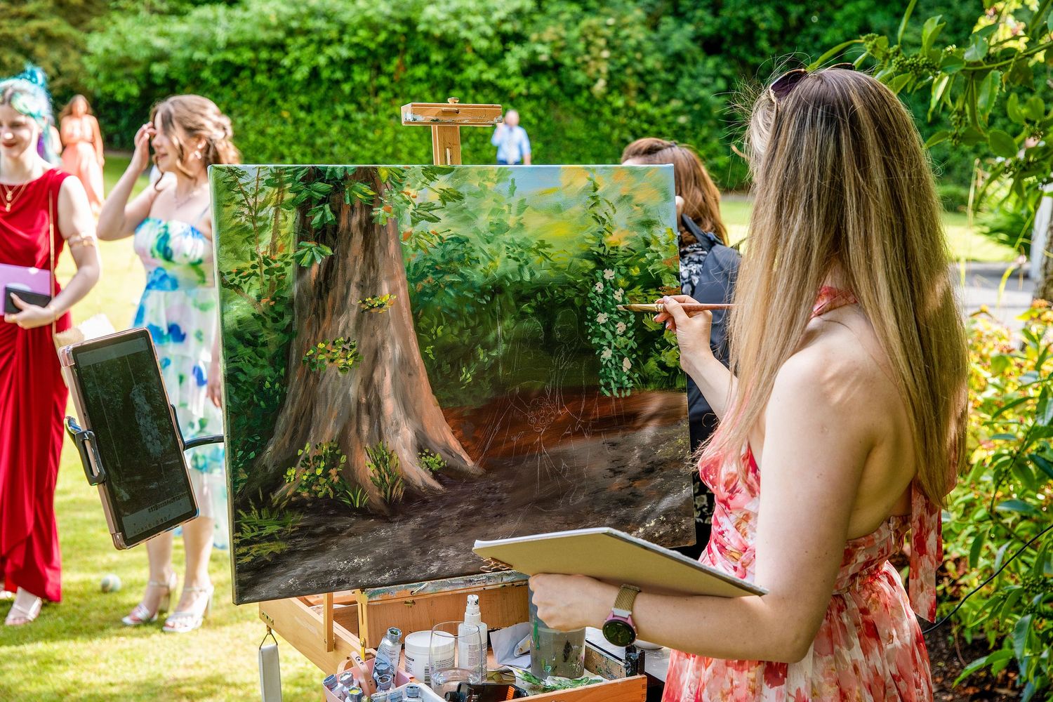 Artist painting a detailed landscape on canvas at an outdoor garden event with guests in formal attire nearby.