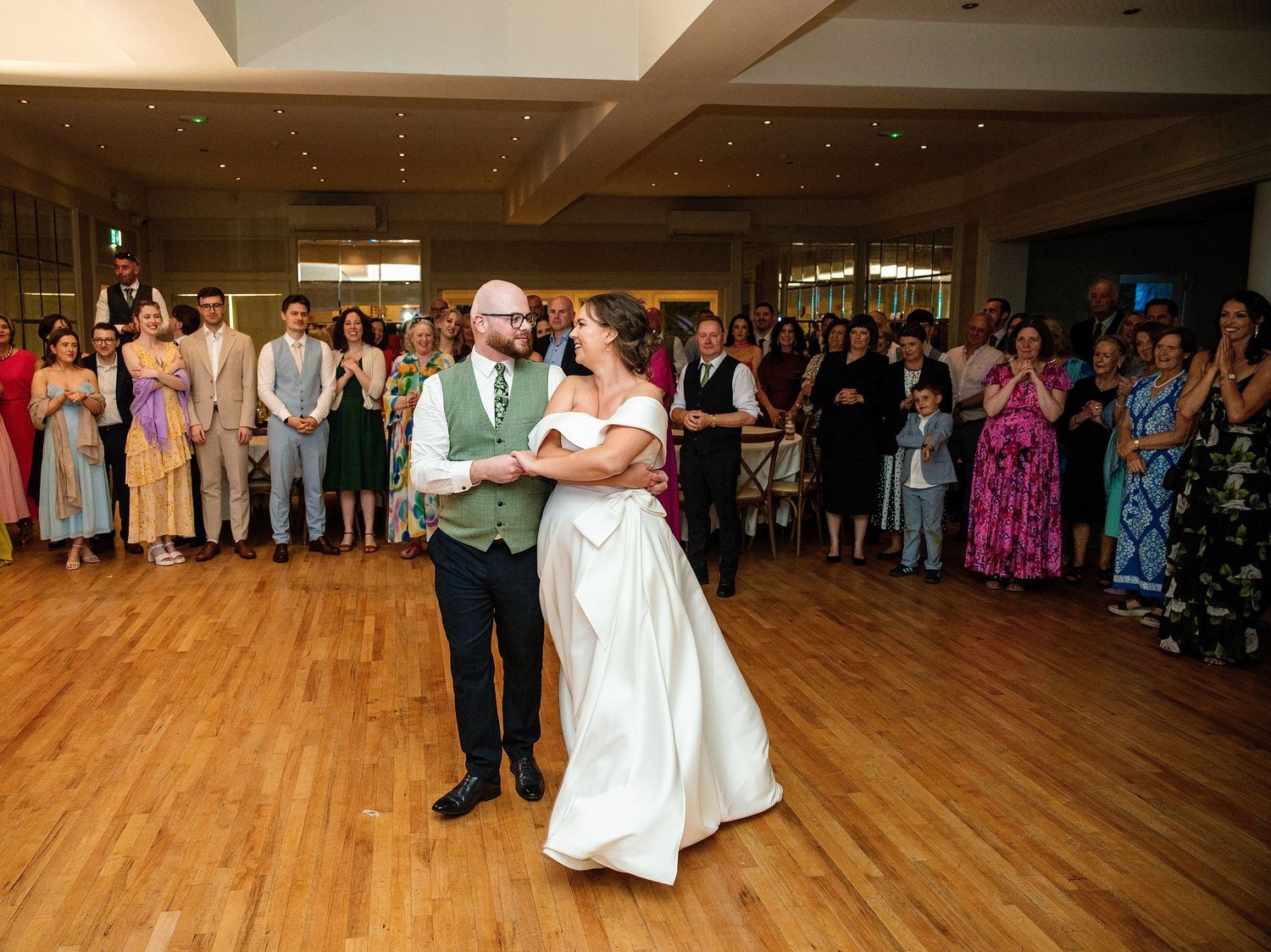 Bride and groom share their first dance on a wooden floor surrounded by smiling wedding guests in a elegant reception hall.