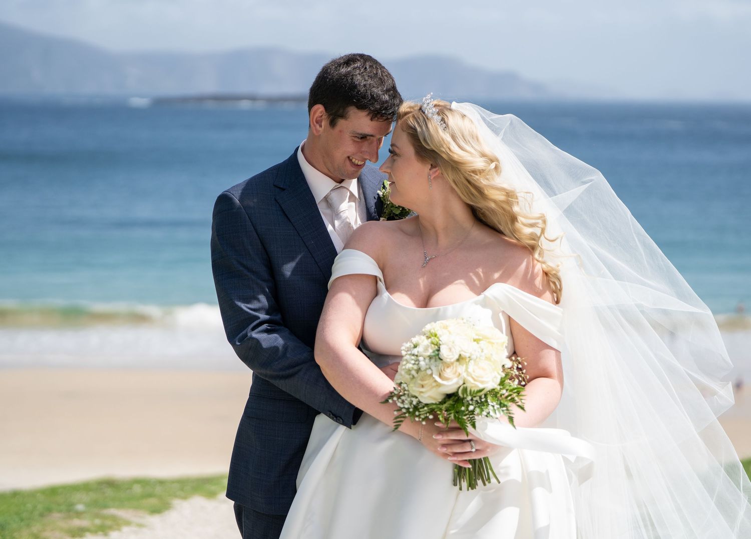 Bride and groom share romantic moment on beach, bride in off-shoulder gown holding white bouquet with veil flowing.