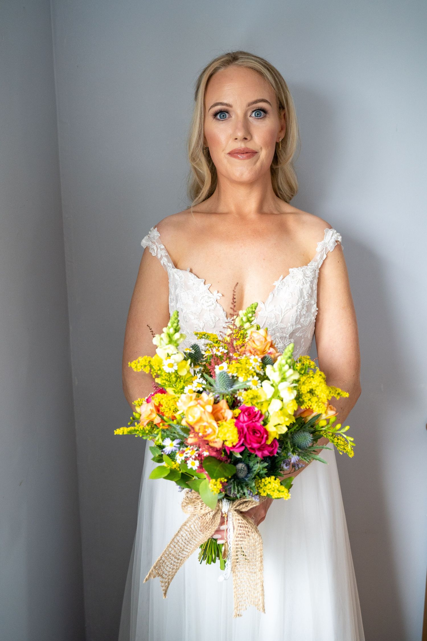 Blonde bride in white lace gown holding colorful yellow, pink, and orange wildflower bouquet with burlap ribbon.