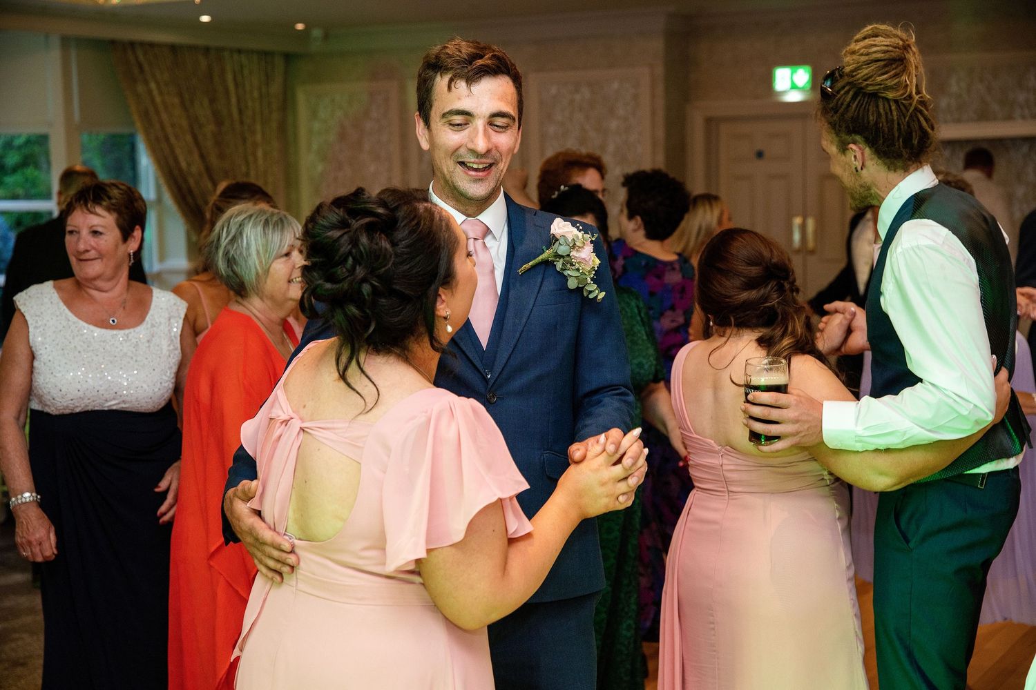 Guests dancing joyfully at a wedding reception, with a smiling groom in a blue suit holding a bouquet.