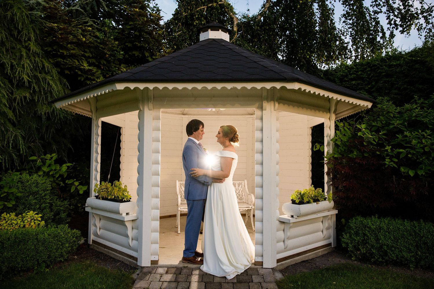 Bride and groom embracing in a white wooden gazebo surrounded by lush greenery during an outdoor wedding ceremony.