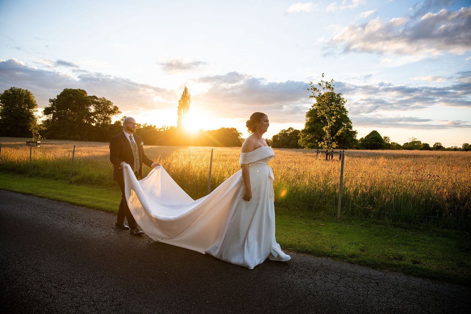 Bride and groom walk at sunset on a country road, her white gown trailing behind them.