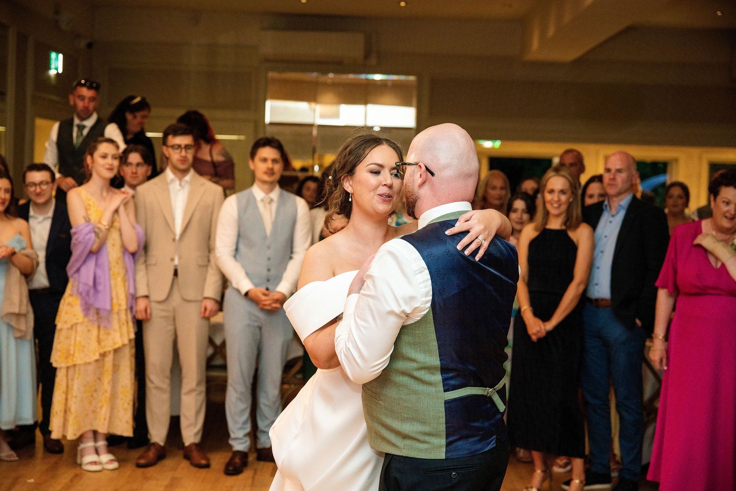 Bride and groom share their first dance surrounded by smiling wedding guests at elegant reception venue.