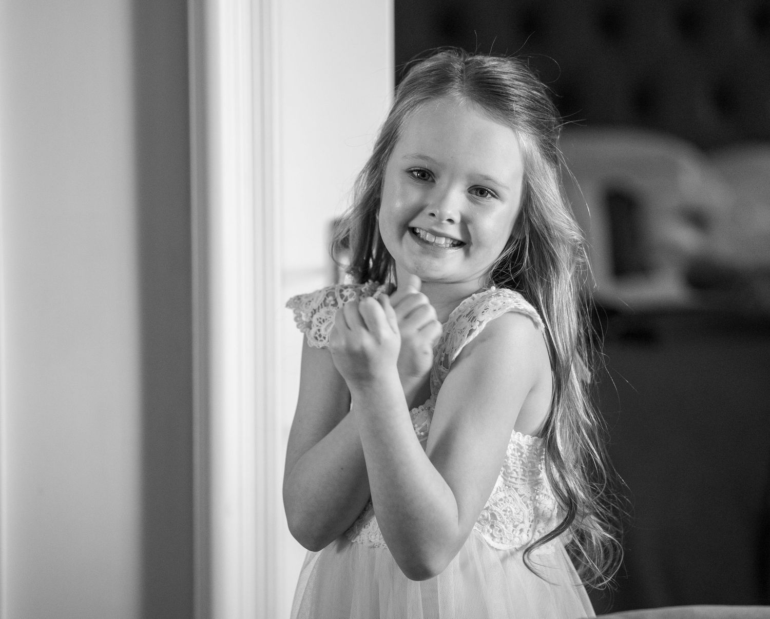 A smiling young girl in a white lace dress poses cheerfully in a black and white portrait photo.