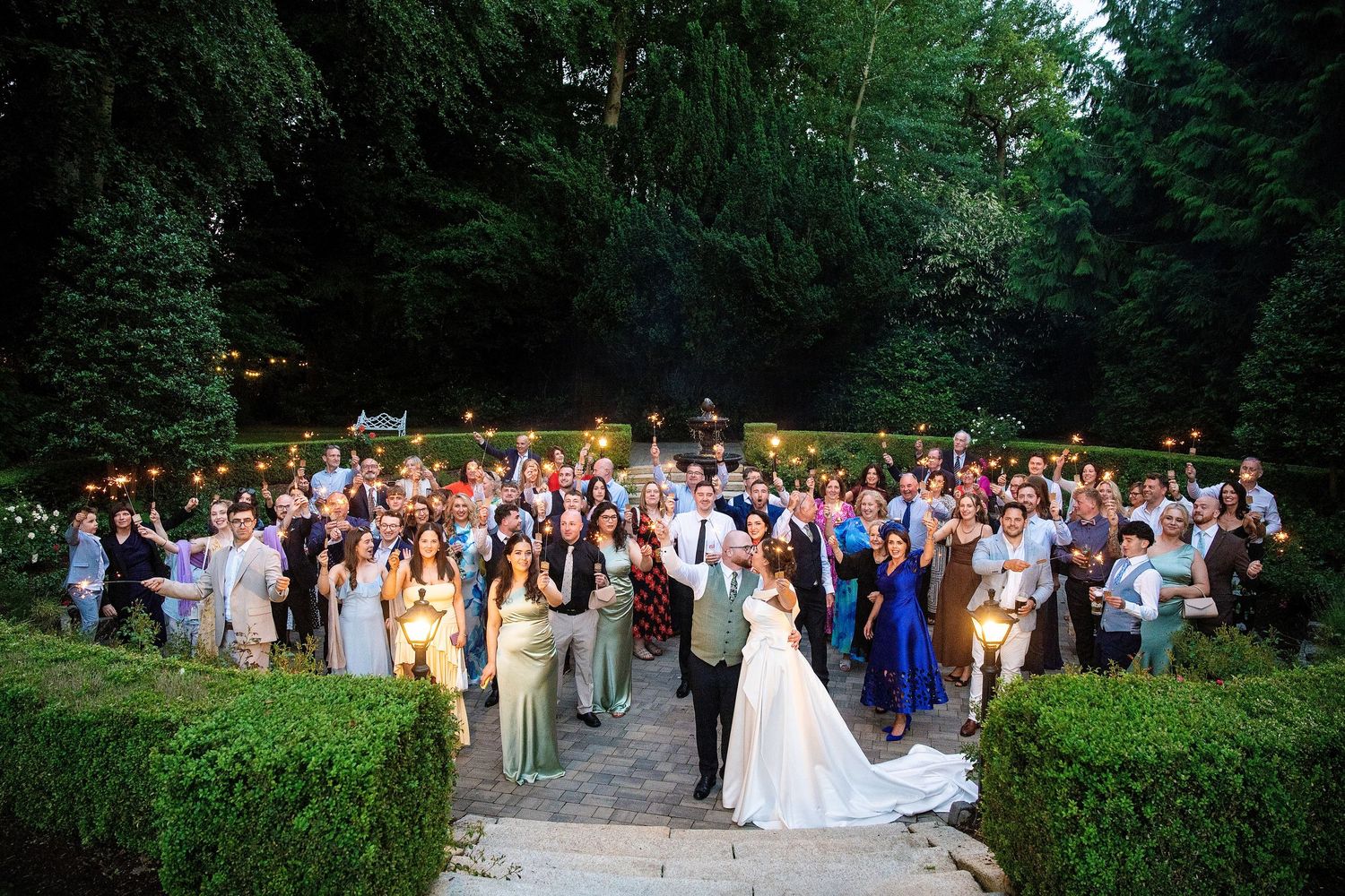 Large wedding party group photo outdoors at dusk, bride and groom centered on stone steps surrounded by guests.