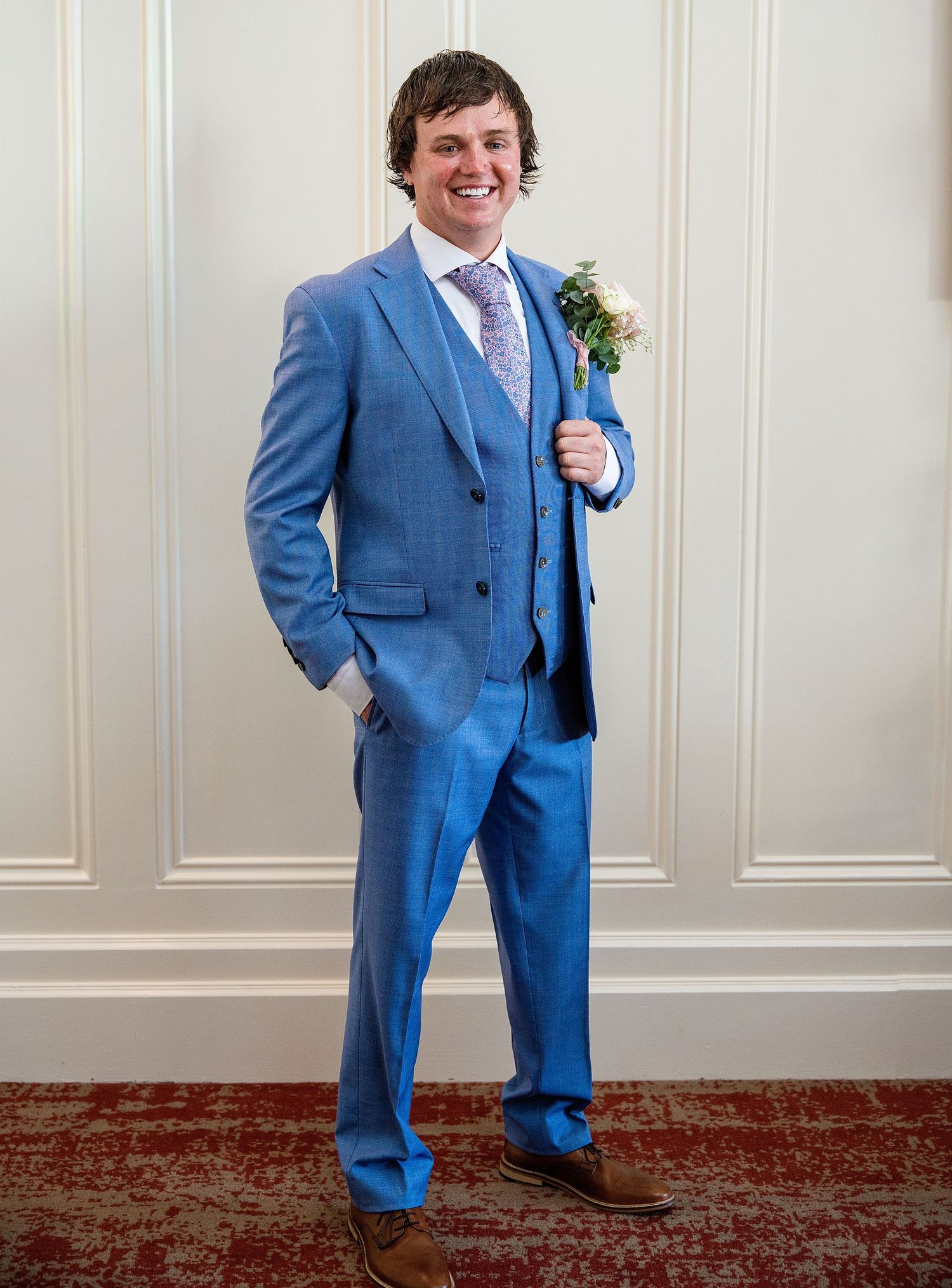 Young man smiling in a blue three-piece suit with floral boutonniere, posing at a formal wedding event.