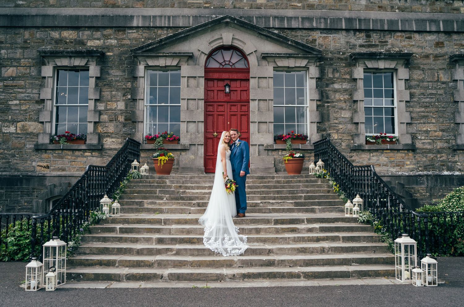 Bride and groom pose on stone steps before a historic building with a red door, flanked by lanterns.