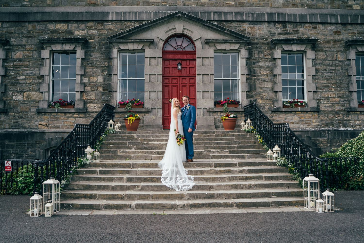 Bride and groom stand on stone steps of elegant historic venue with red door and lanterns at Bellinter house.