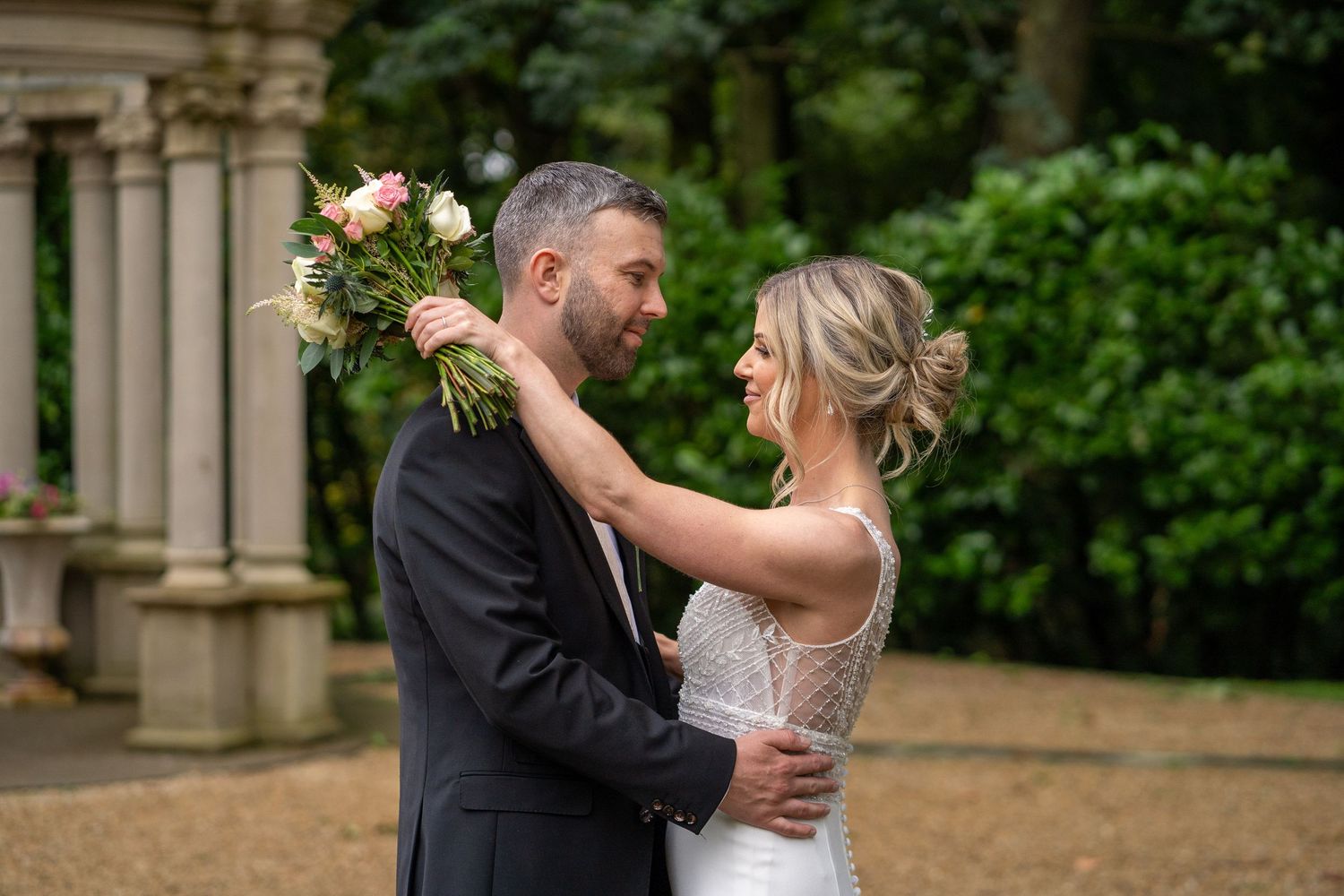 Newlywed couple shares romantic moments in a garden setting with stone columns and lush greenery.