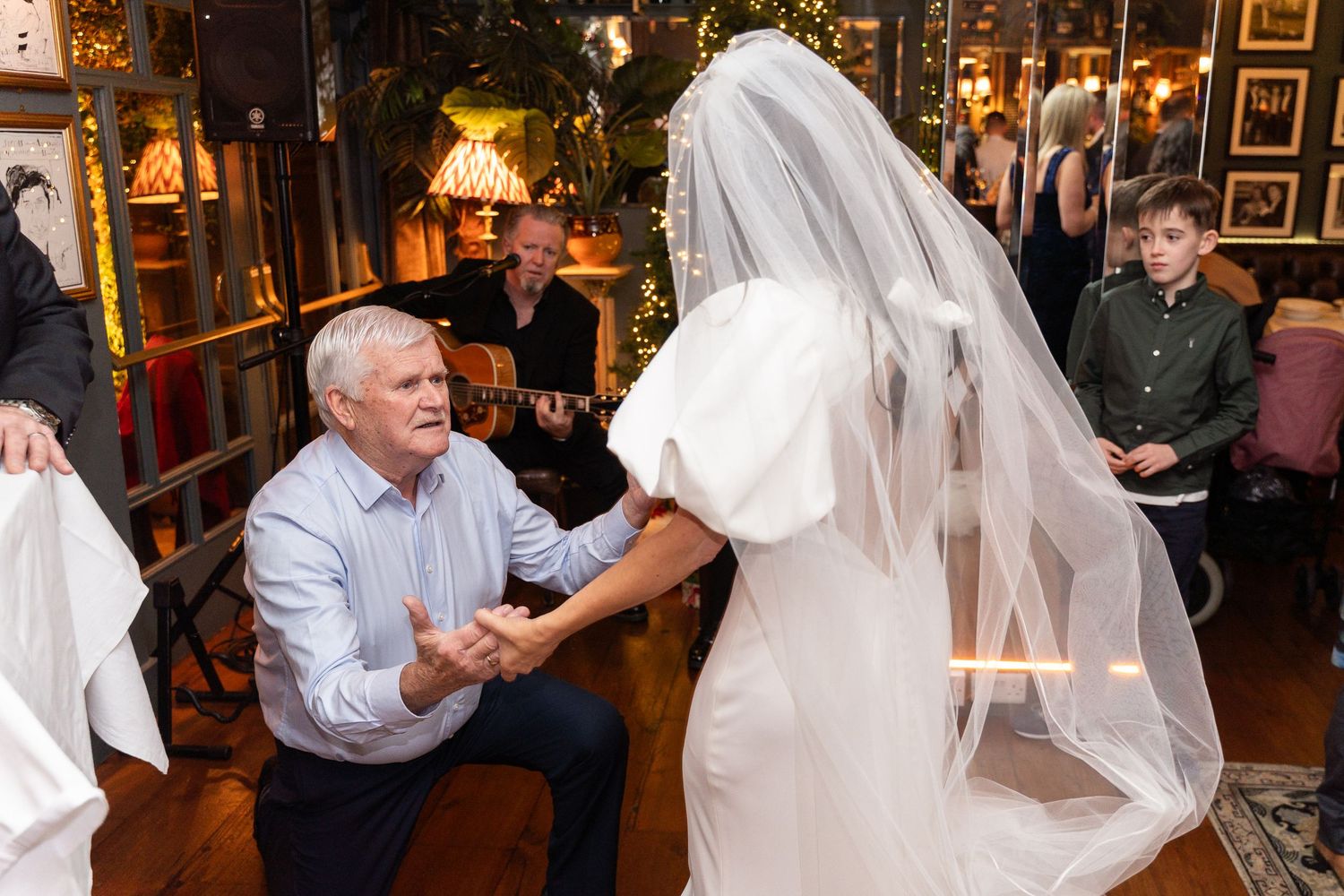 Elderly man kneeling before a bride in a white wedding dress and veil during a wedding reception dance.