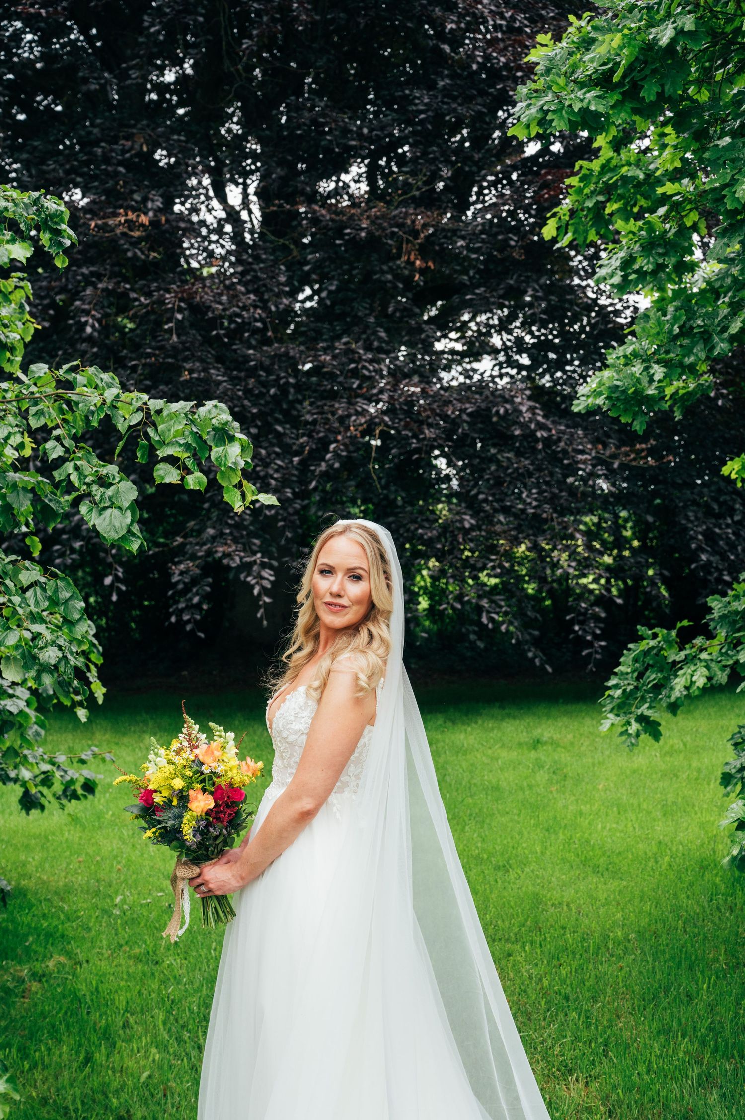 Bride in white gown and veil holding colorful bouquet standing in lush green garden with dark foliage backdrop at Bellinter house.